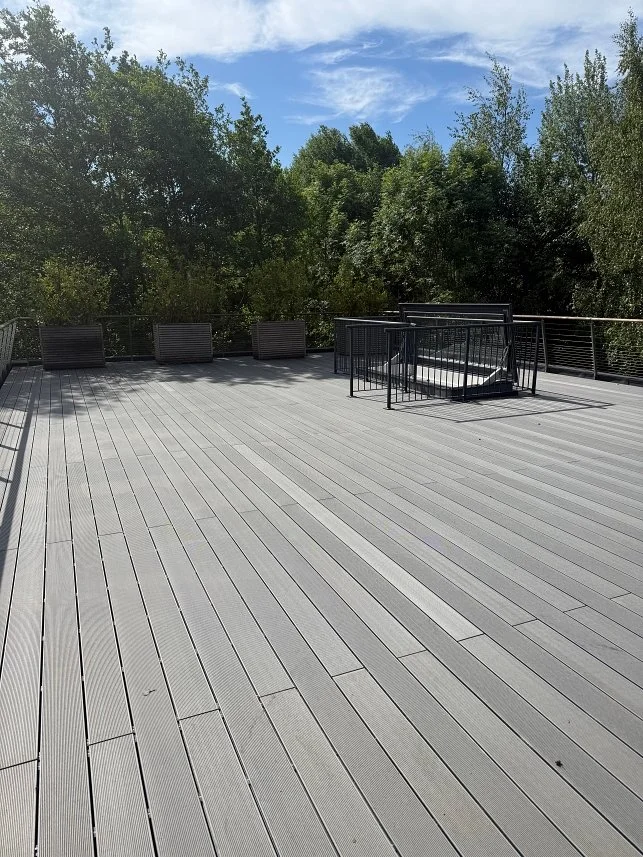 A spacious outdoor wooden deck with black chairs along the edge, surrounded by lush green trees under a partly cloudy sky.