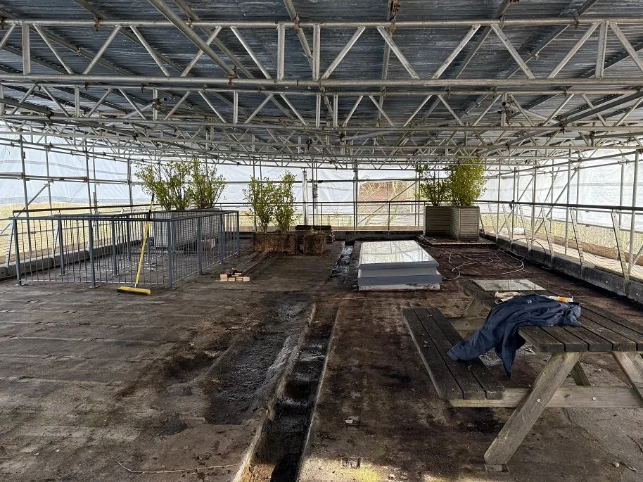 Interior view of a greenhouse under construction or renovation, with exposed metal framework, dusty floor, and some potted plants. A broom is leaning against a railing, and there's a picnic table with a jacket on it.