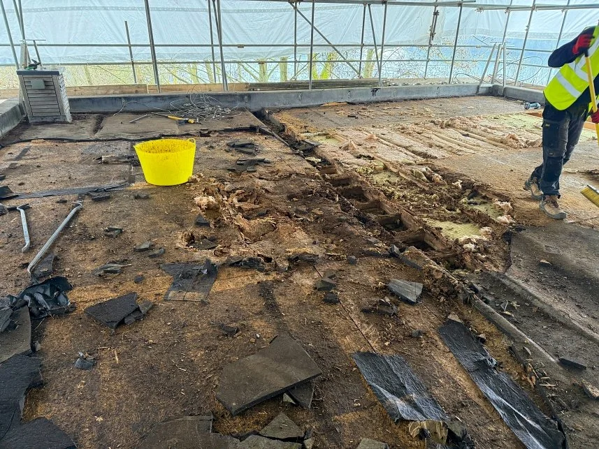 Remains of a demolished or damaged floor with scattered debris inside a greenhouse or similar structure. A worker wearing a safety vest and gloves is partially visible on the right.