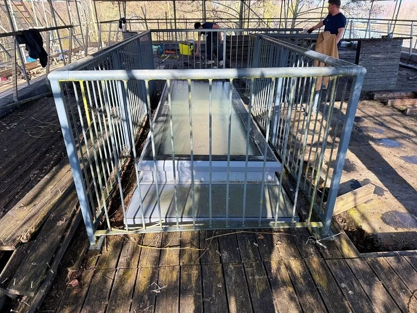 Metal cage on a wooden deck with two people in the background, one standing and the other sitting, near tools and supplies.