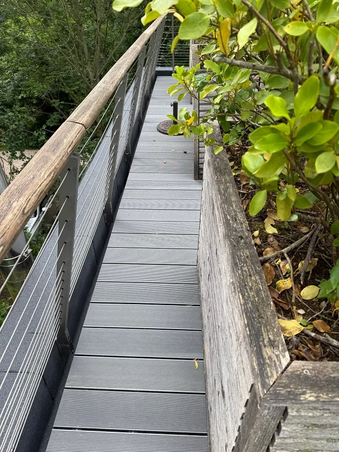 Narrow outdoor wooden bridge with gray composite decking, metal railing, and a wooden handrail, surrounded by green foliage.