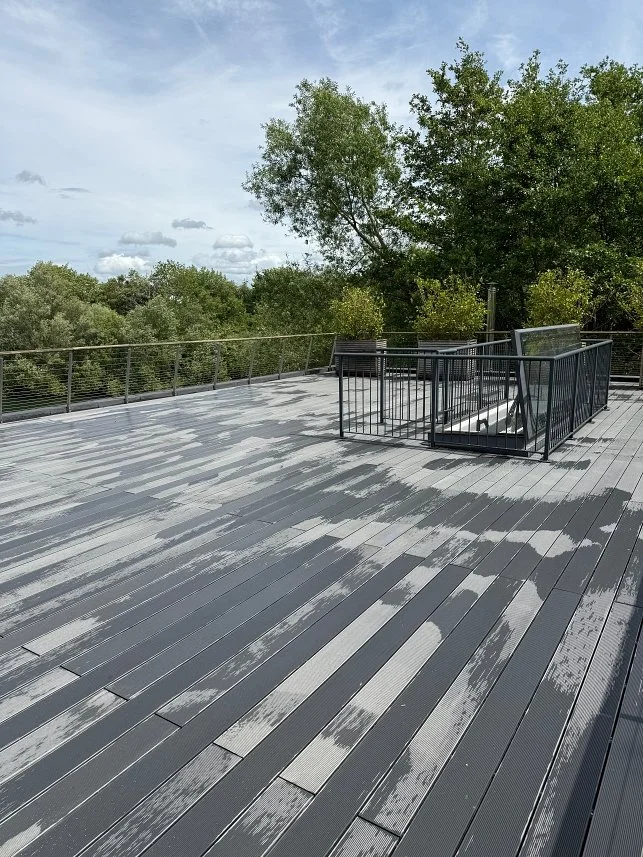 Empty outdoor deck with black railing, surrounded by green trees and a partly cloudy sky.