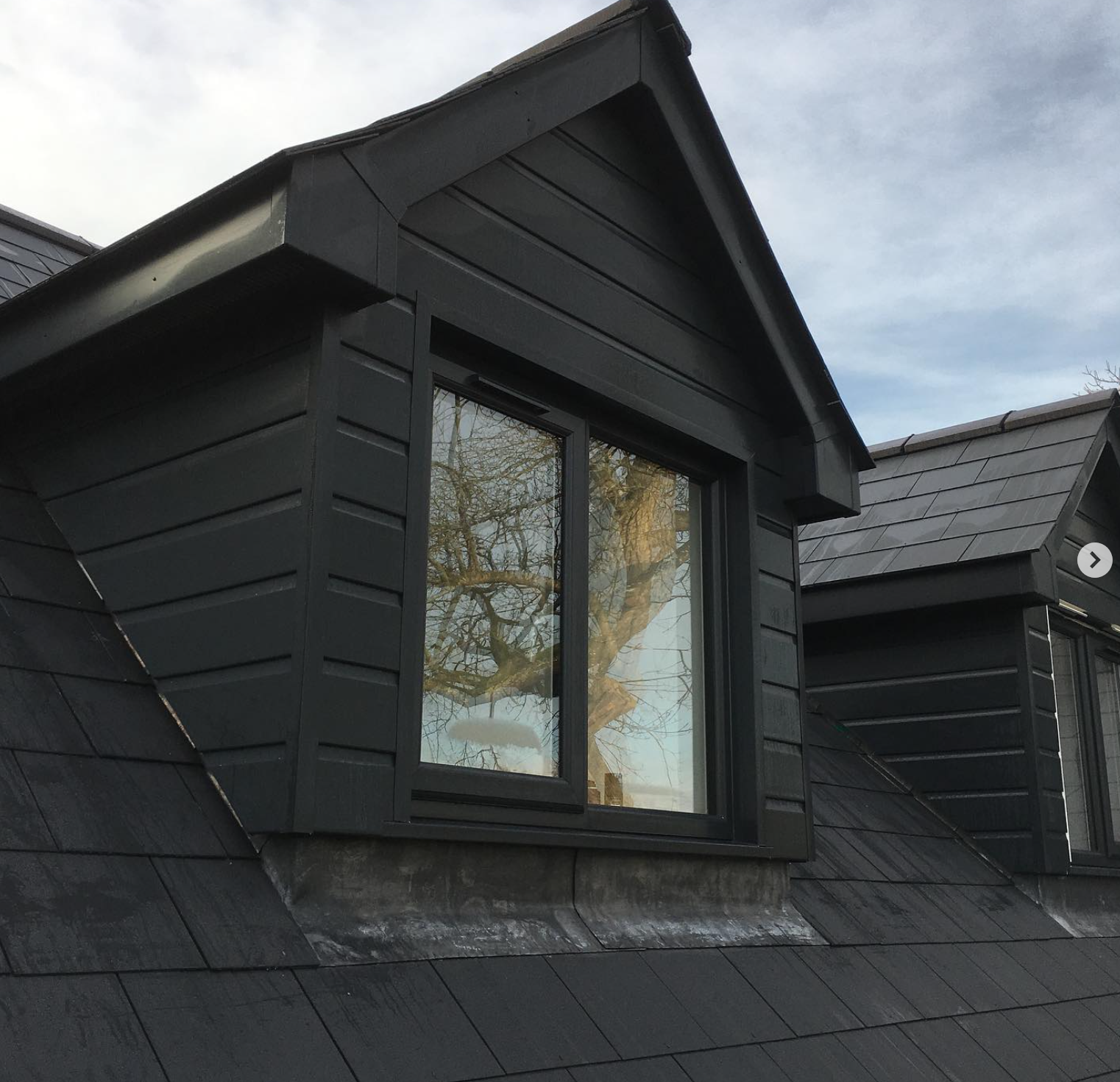 Close-up of a black gable roof dormer window on a dark house, with a reflection of a leafless tree in the glass.