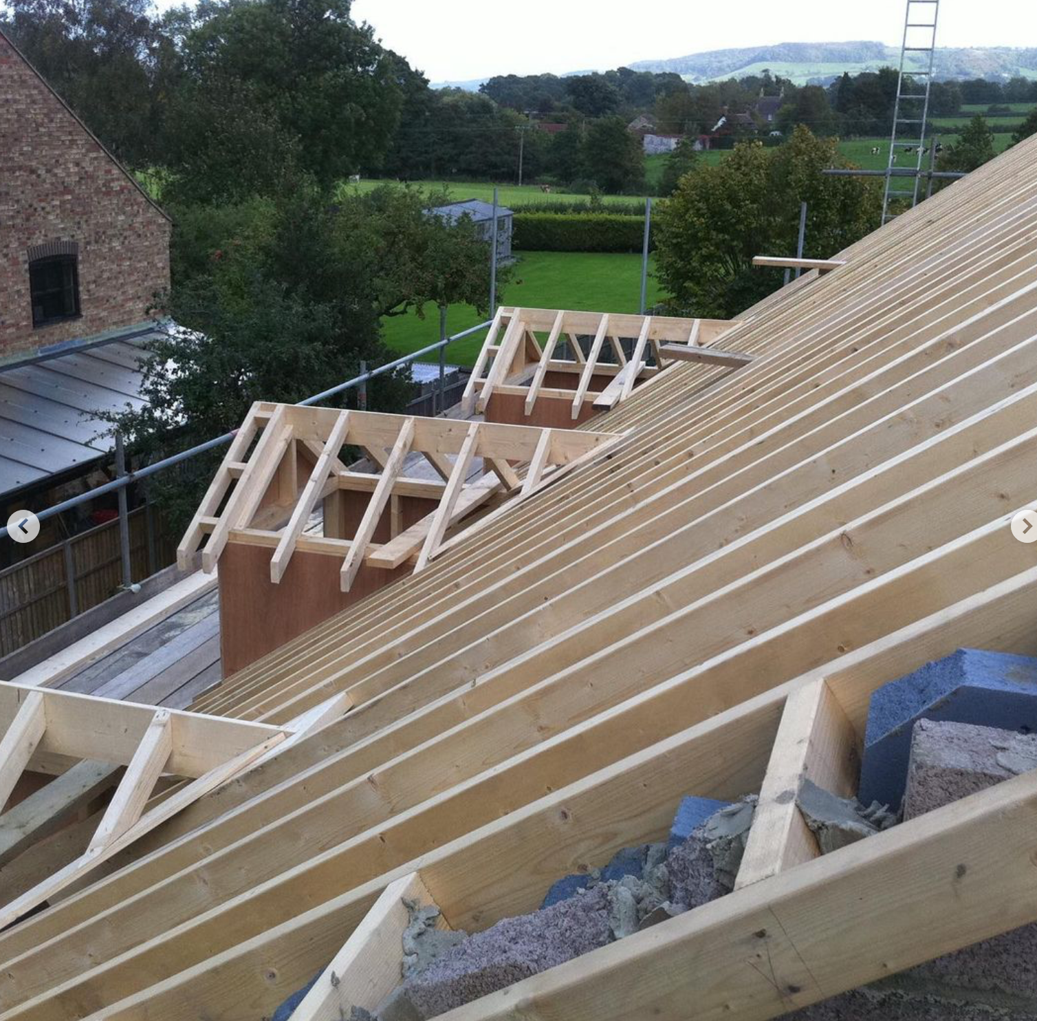 Wooden roof framing under construction on a residential building with a scenic backyard and distant hills in the background.