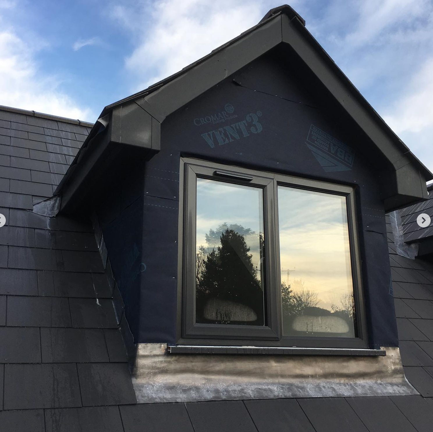 Close-up of a newly installed dormer window on a roof, with black siding and reflection of sky and trees in the window glass.