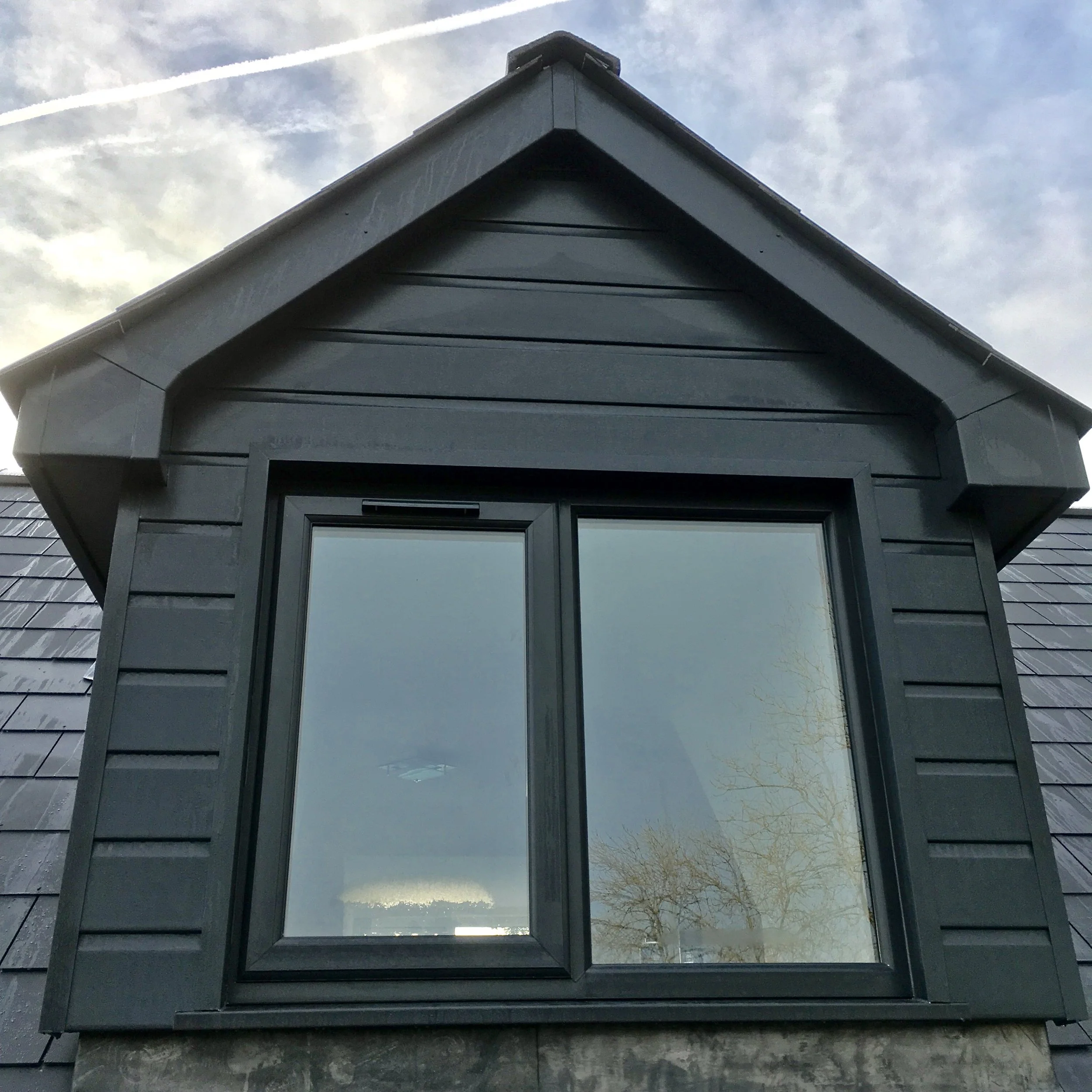 Close-up of a black-framed dormer window on a dark grey house with shingled siding, under a cloudy sky.