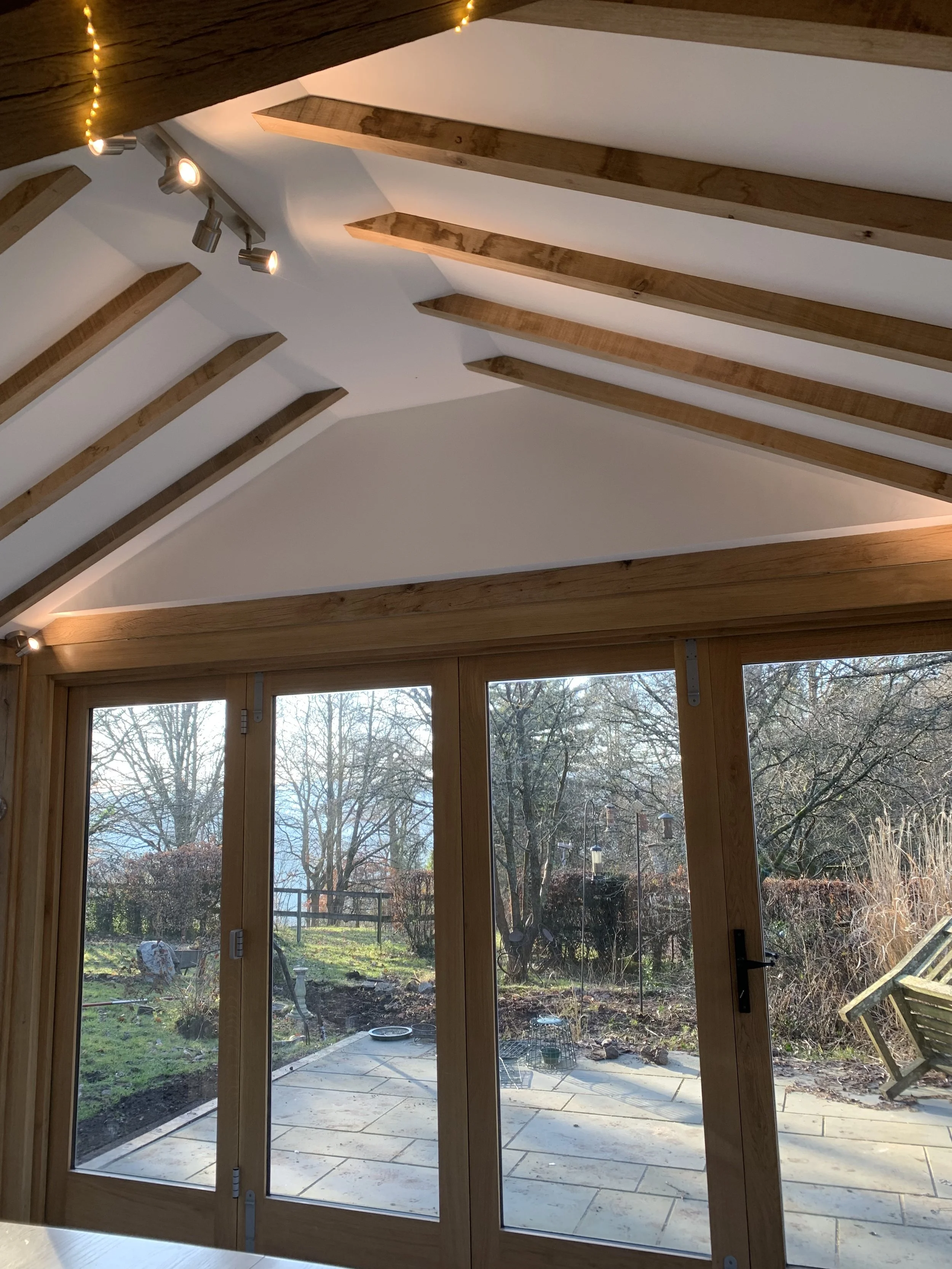 Interior view of a room with a vaulted ceiling featuring exposed wooden beams, track lighting, and large glass sliding doors opening to a backyard with trees and outdoor furniture.