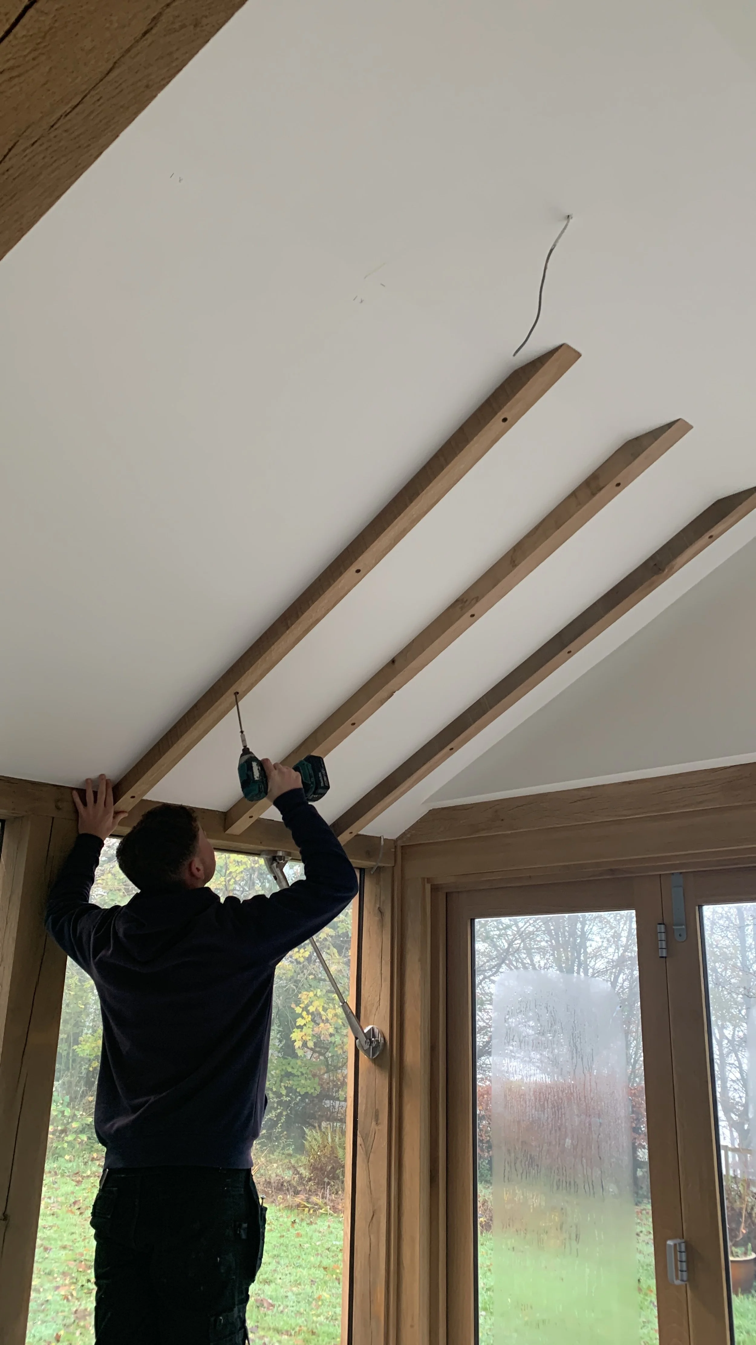A man installing wooden beams on a sloped ceiling with a cordless drill inside a room with large windows.