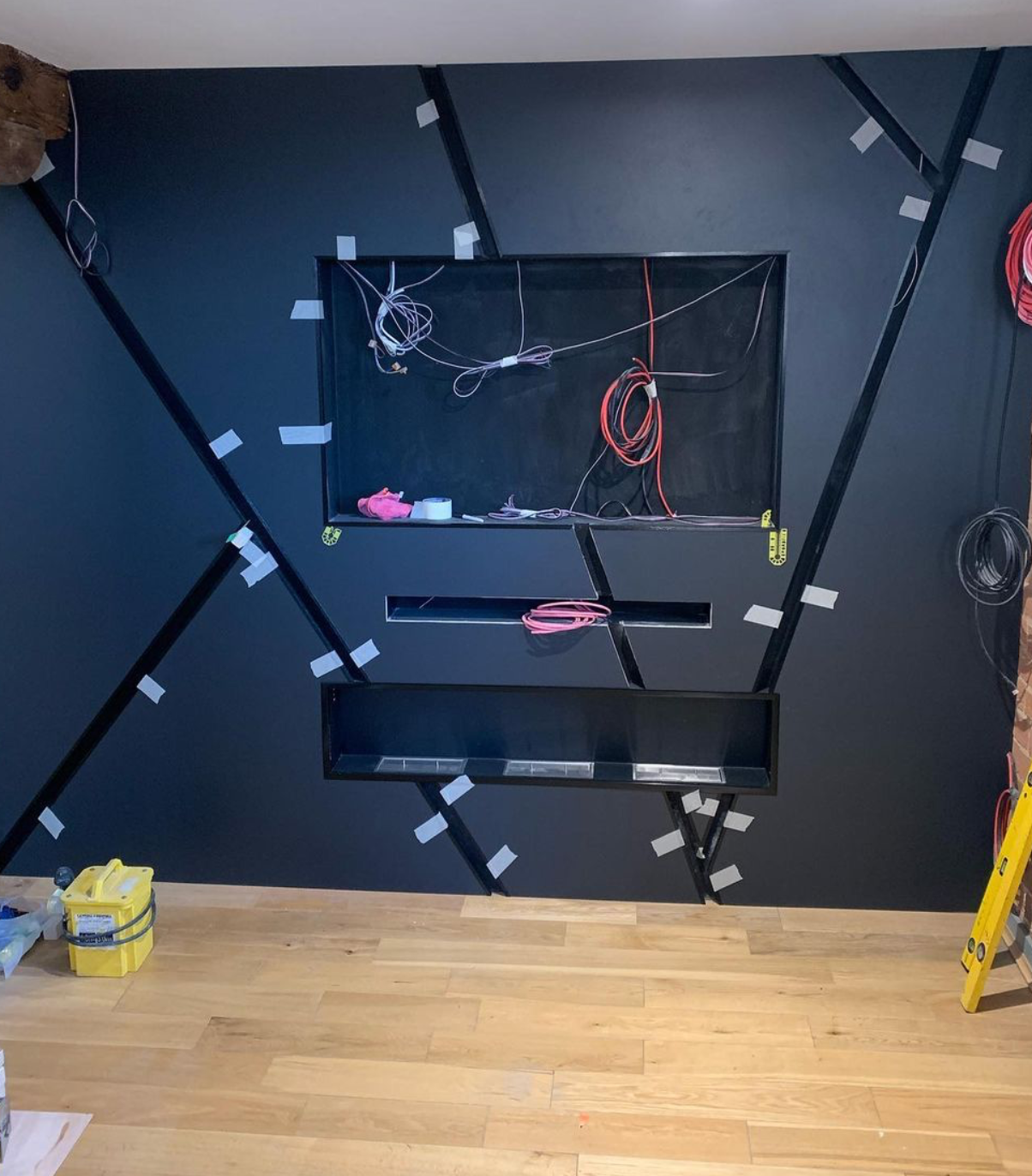 Dark wall with geometric black frames, wires hanging, and some tools and materials on floor and wall, indicating ongoing construction or installation.