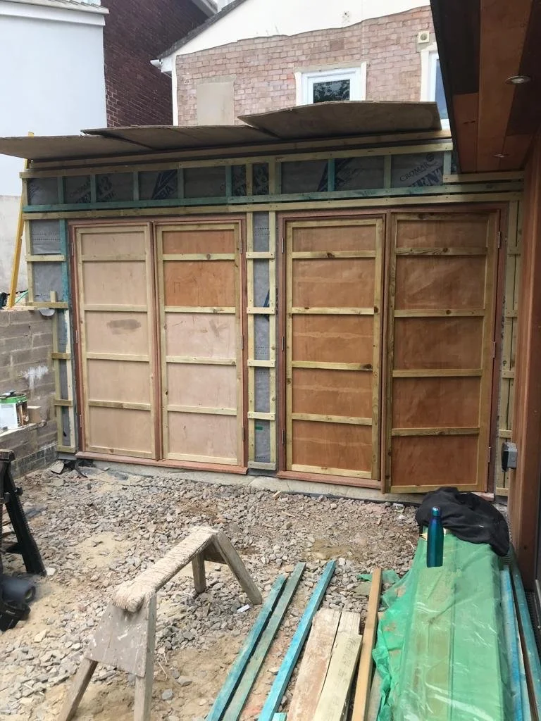 Construction site with wooden framing and plywood panels, partially built wall, gravel ground, construction tools and materials, adjacent brick building and house with window in background.