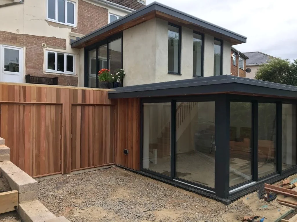 Newly constructed modern extension with large glass windows and black frames, adjacent to a brick building, in a backyard under construction, with a wooden fence and some potted plants on the upper level.