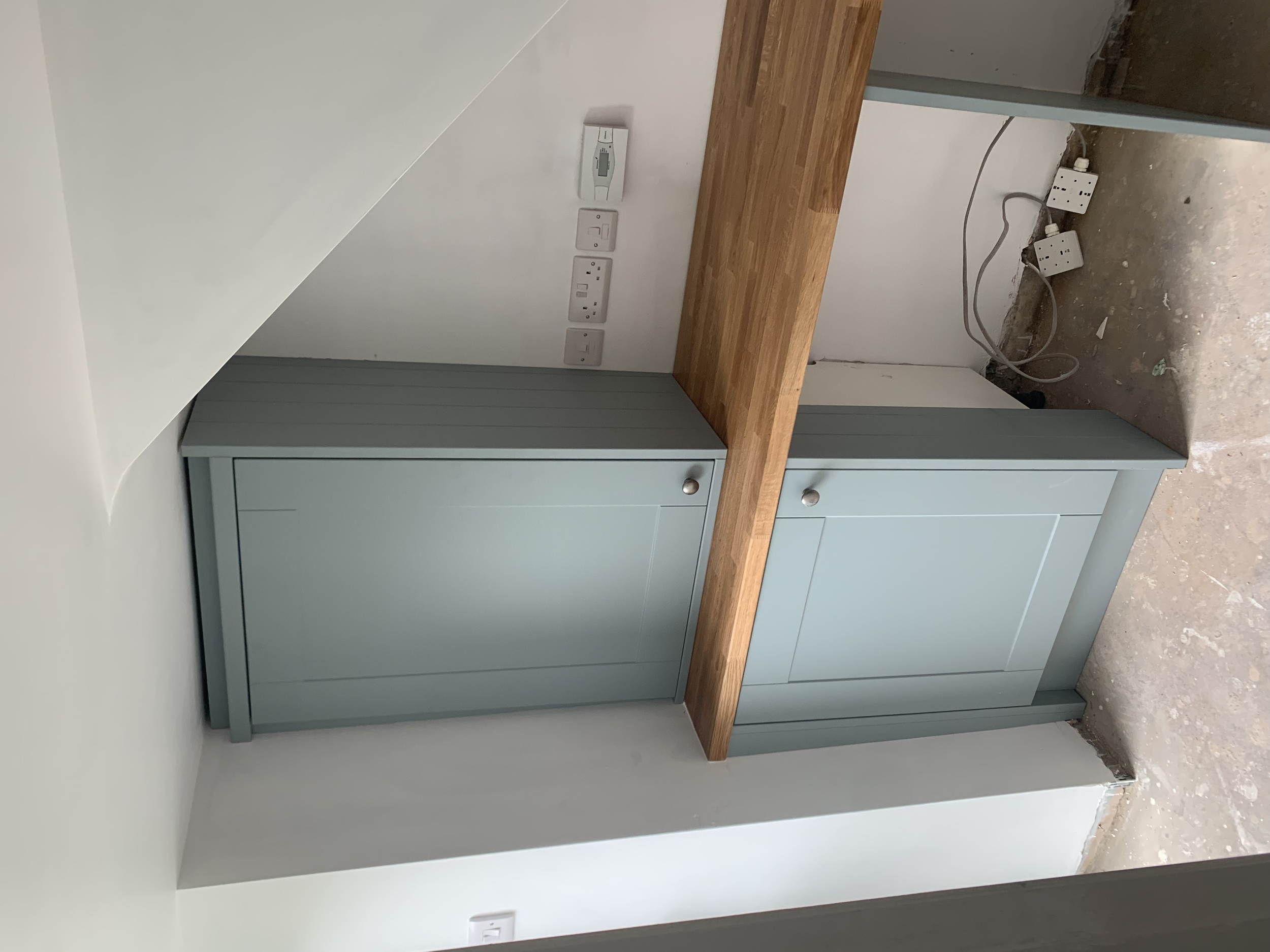 Oak kitchen countertop with light blue cabinets beneath, white wall with electrical outlets and switches, unfinished floor with visible wires and sockets.