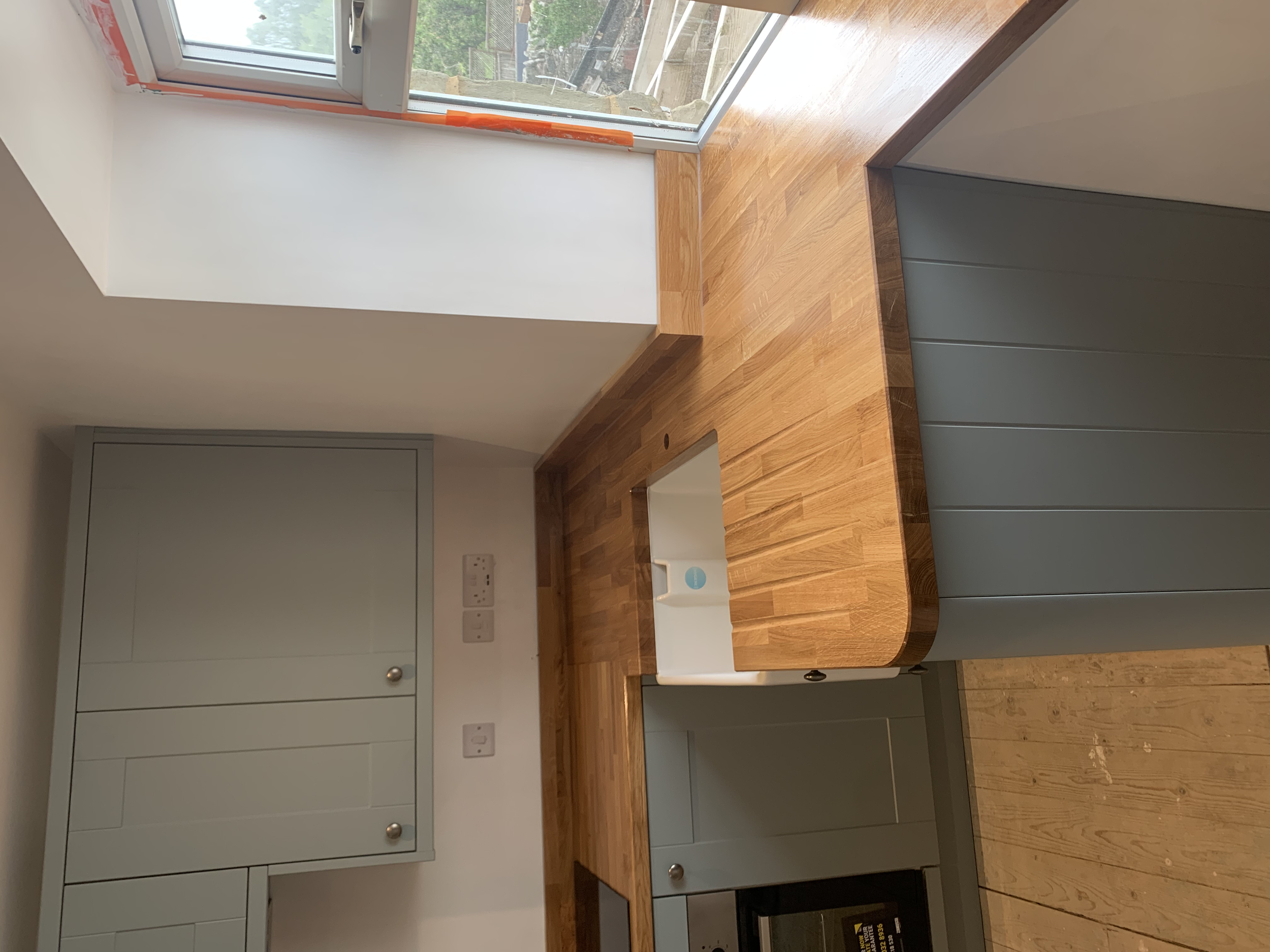 A modern kitchen with light green cabinets, a wooden countertop, and a farmhouse sink. There is a large window above the sink, allowing natural light into the space.