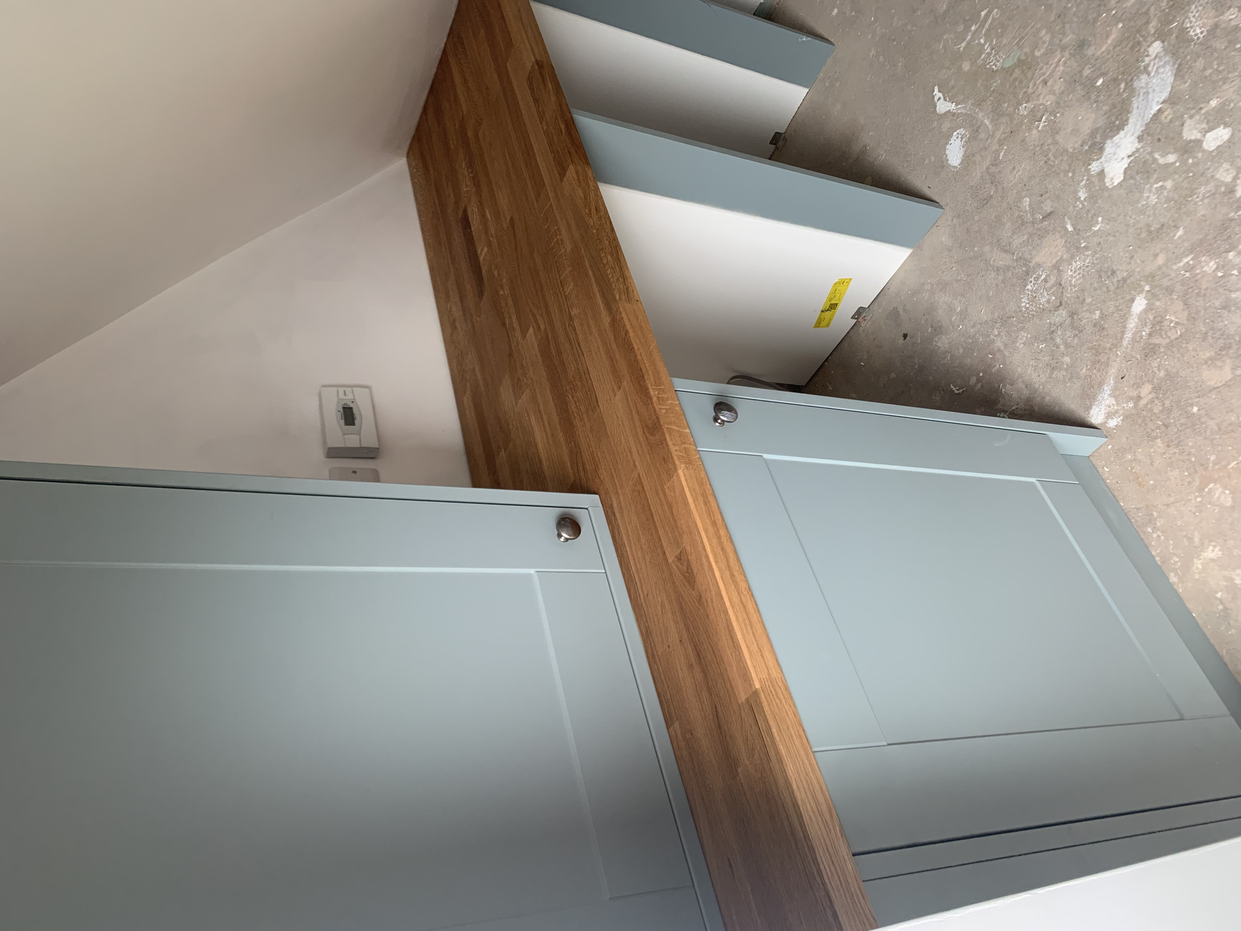 Empty light blue kitchen cabinet with a wooden countertop, under construction or renovation, with a power outlet on the white wall and a concrete floor with paint splatters.