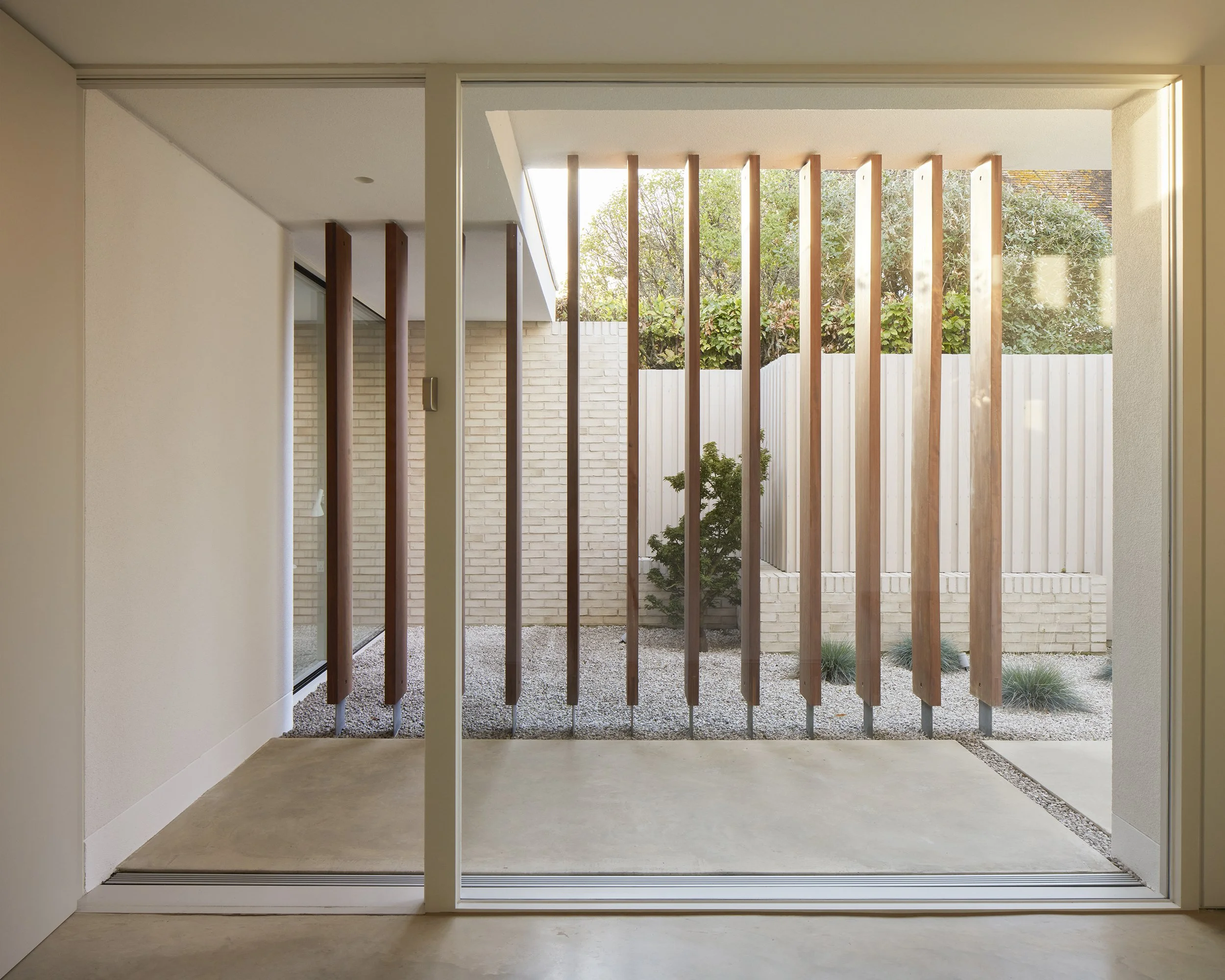 Empty modern patio with wooden vertical slats, small plants, and a white fence, viewed through sliding glass doors from inside.