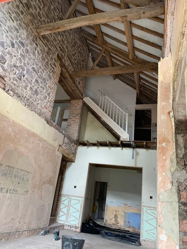 Interior of a building under renovation with exposed brick walls, wooden roof beams, and an upstairs staircase with white railings.