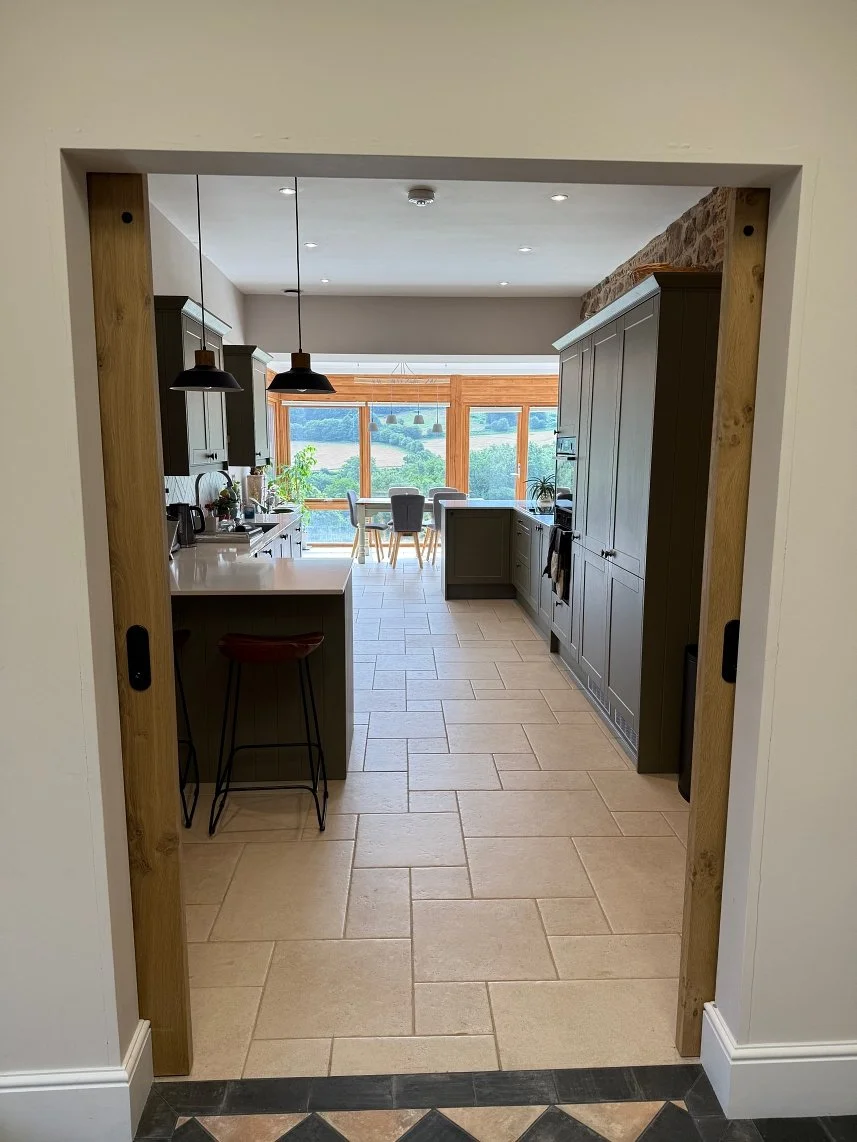 View of a modern kitchen with dark green cabinets, a white countertop island, and a dining area with large windows overlooking a green landscape.