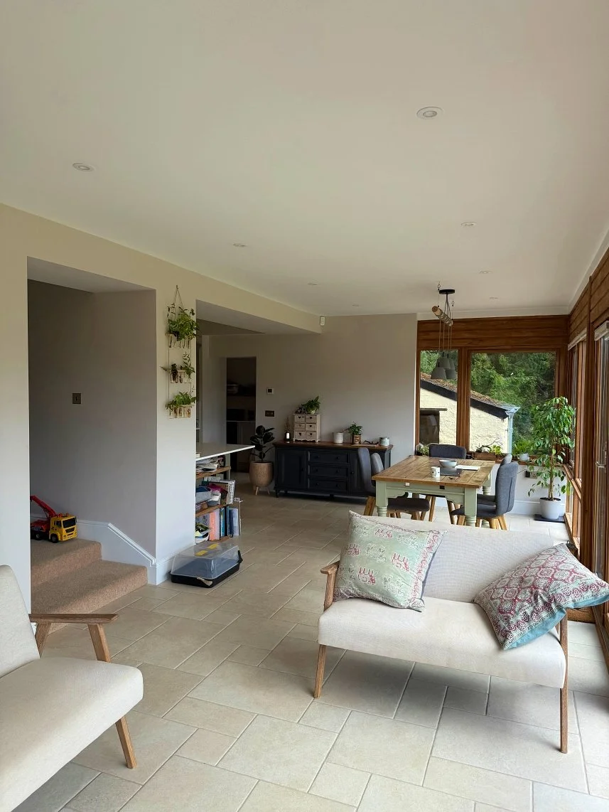 Bright living room with beige tiled floors, a white sofa with patterned pillows, a wooden dining table with four chairs, large windows with wooden frames, and indoor plants.