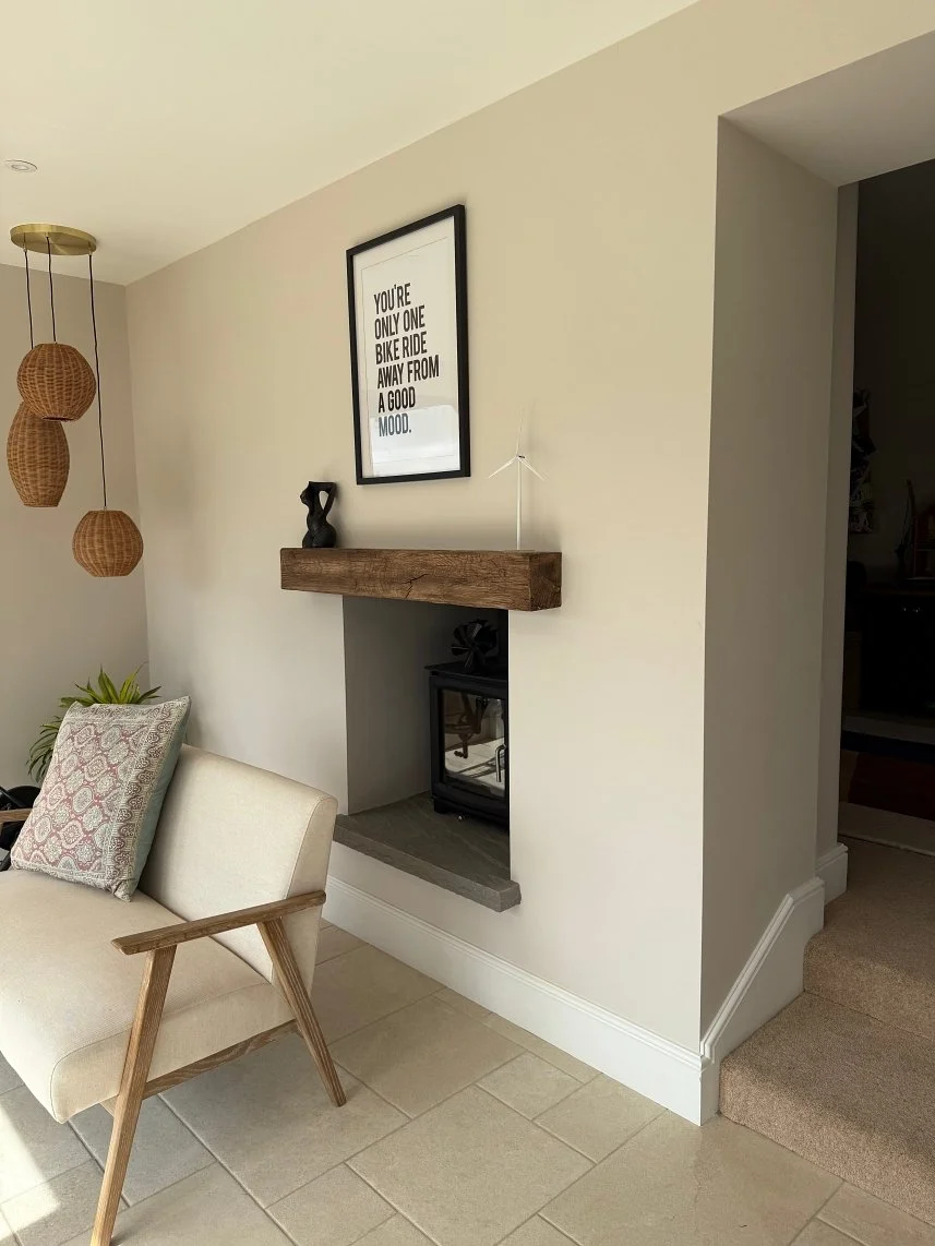 Living room corner with a beige armchair, decorative pillow, green plant, hanging wicker pendant lights, framed quote on wall, small fireplace with black glass door, wooden mantel, and a gray hearth.
