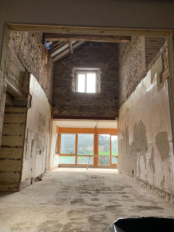 Interior of a building under renovation showing exposed brick walls, unfinished flooring, and a large window with a view of greenery outside.