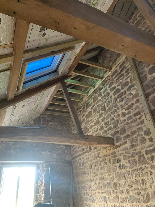 Interior of an unfinished attic with wooden beams, brick walls, a skylight window, and some insulation material.