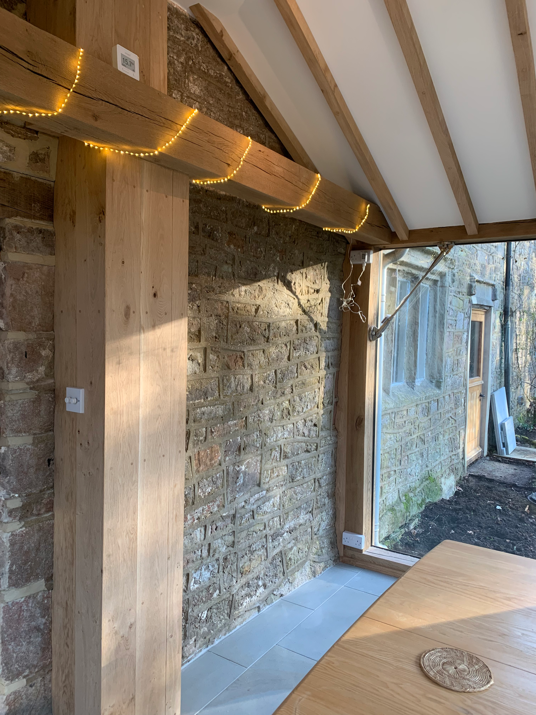 Interior view of a room with a brick wall, wooden beams, a large glass door leading outside, a wooden table, and a brick floor. There are string lights on the wooden beams and an electrical outlet visible on the brick wall.