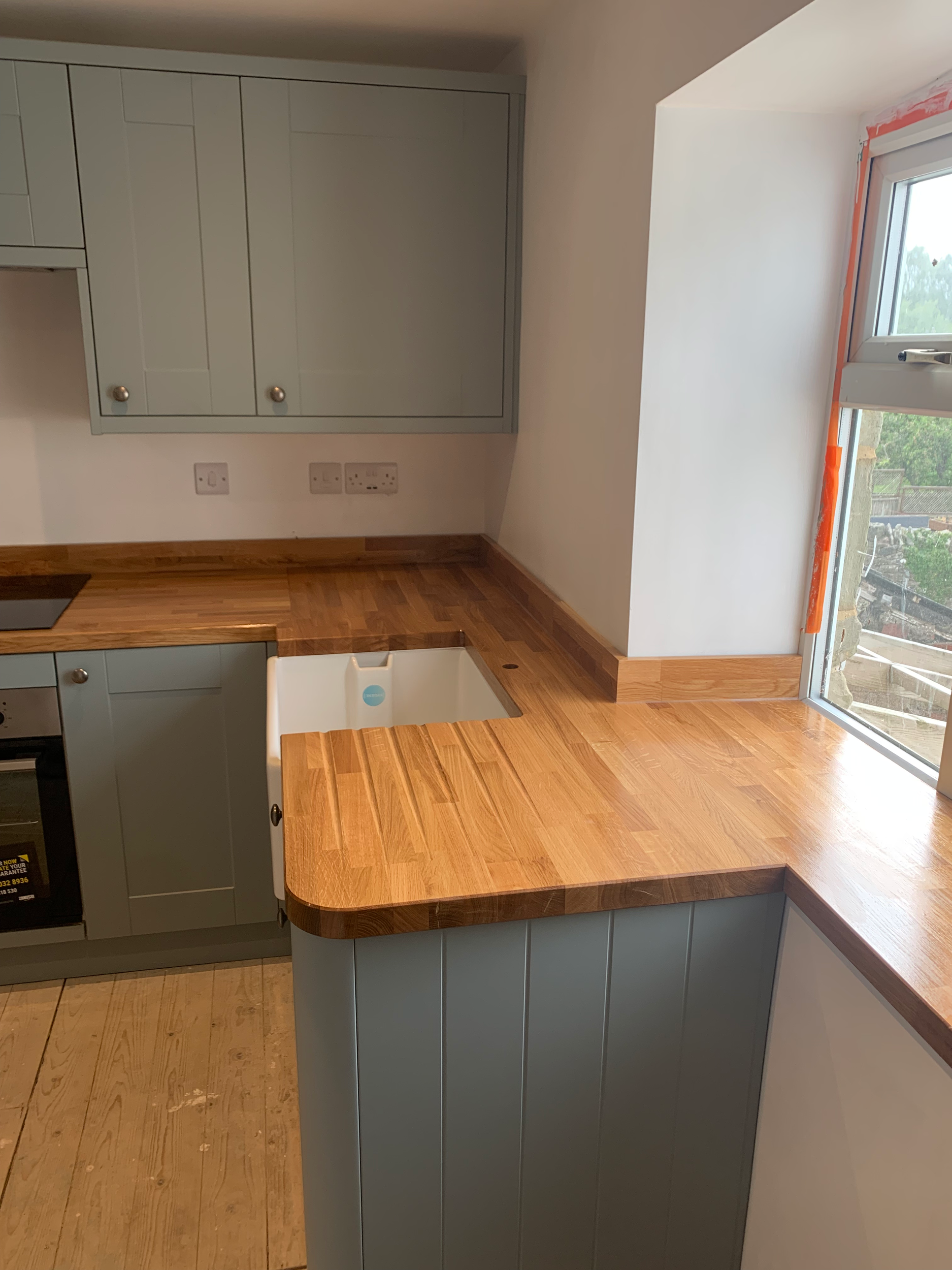 A modern kitchen with light green cabinets, a wooden countertop, and a farmhouse sink. There is a large window above the sink, allowing natural light into the space.