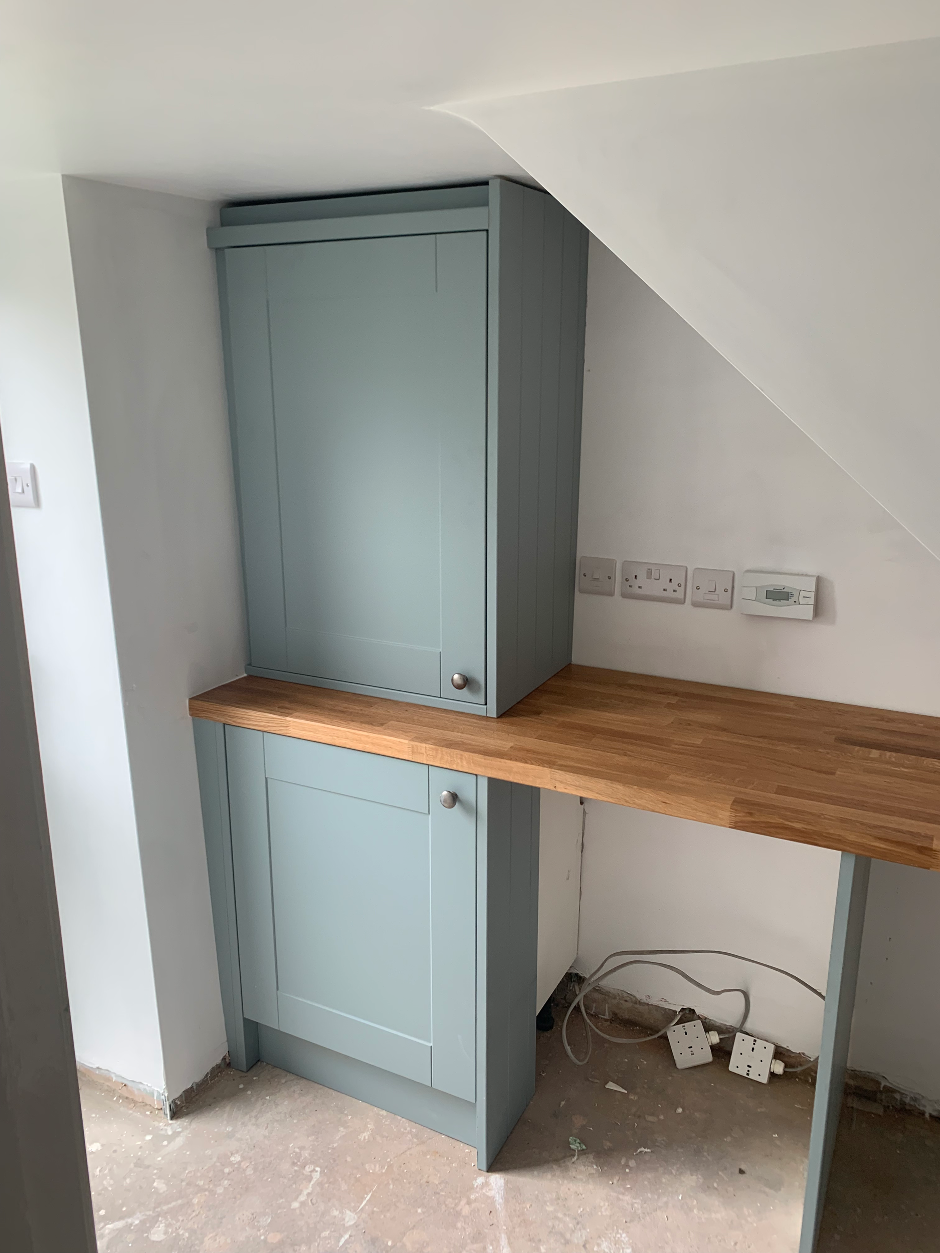 Oak kitchen countertop with light blue cabinets beneath, white wall with electrical outlets and switches, unfinished floor with visible wires and sockets.