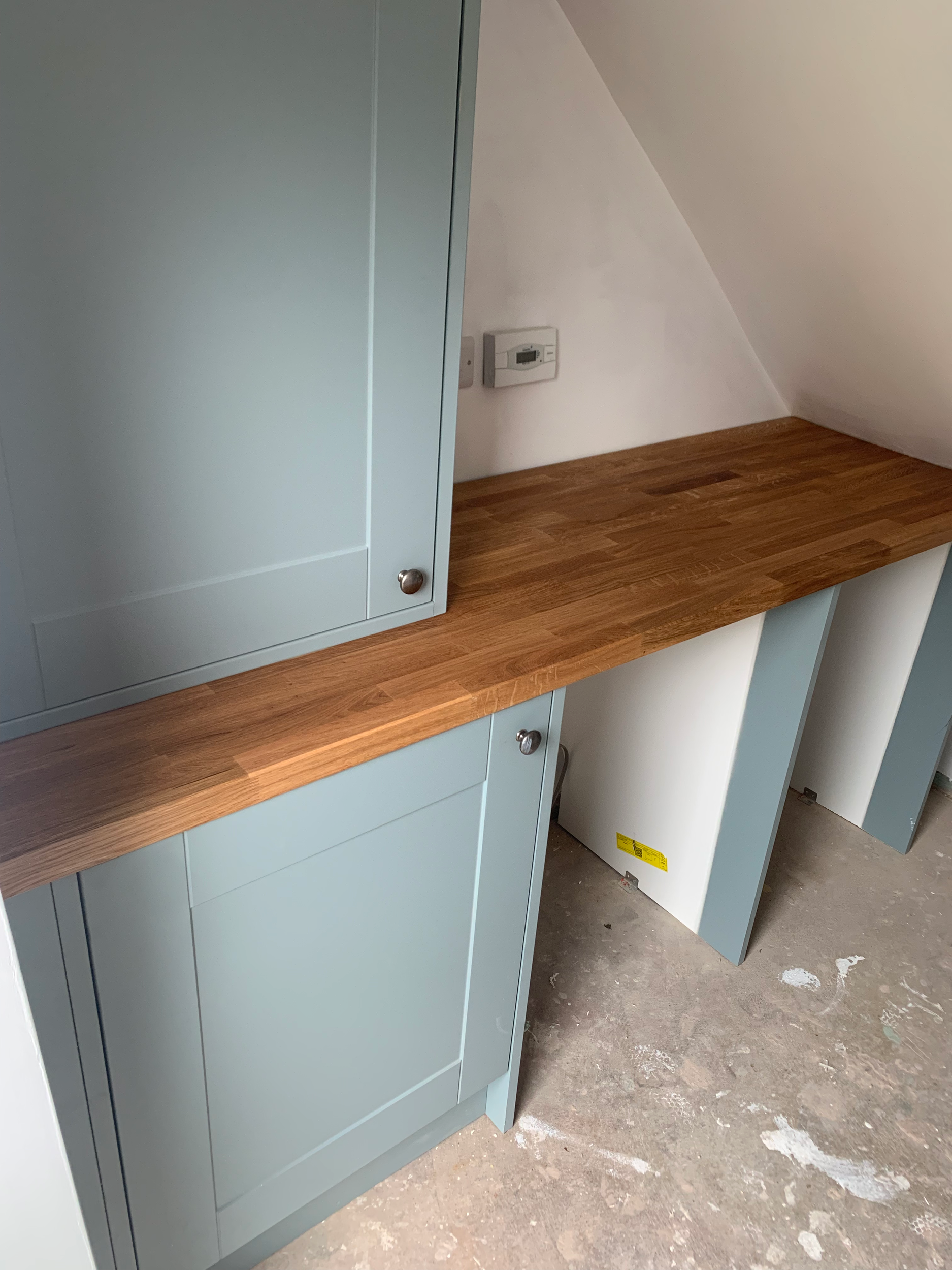 Empty light blue kitchen cabinet with a wooden countertop, under construction or renovation, with a power outlet on the white wall and a concrete floor with paint splatters.