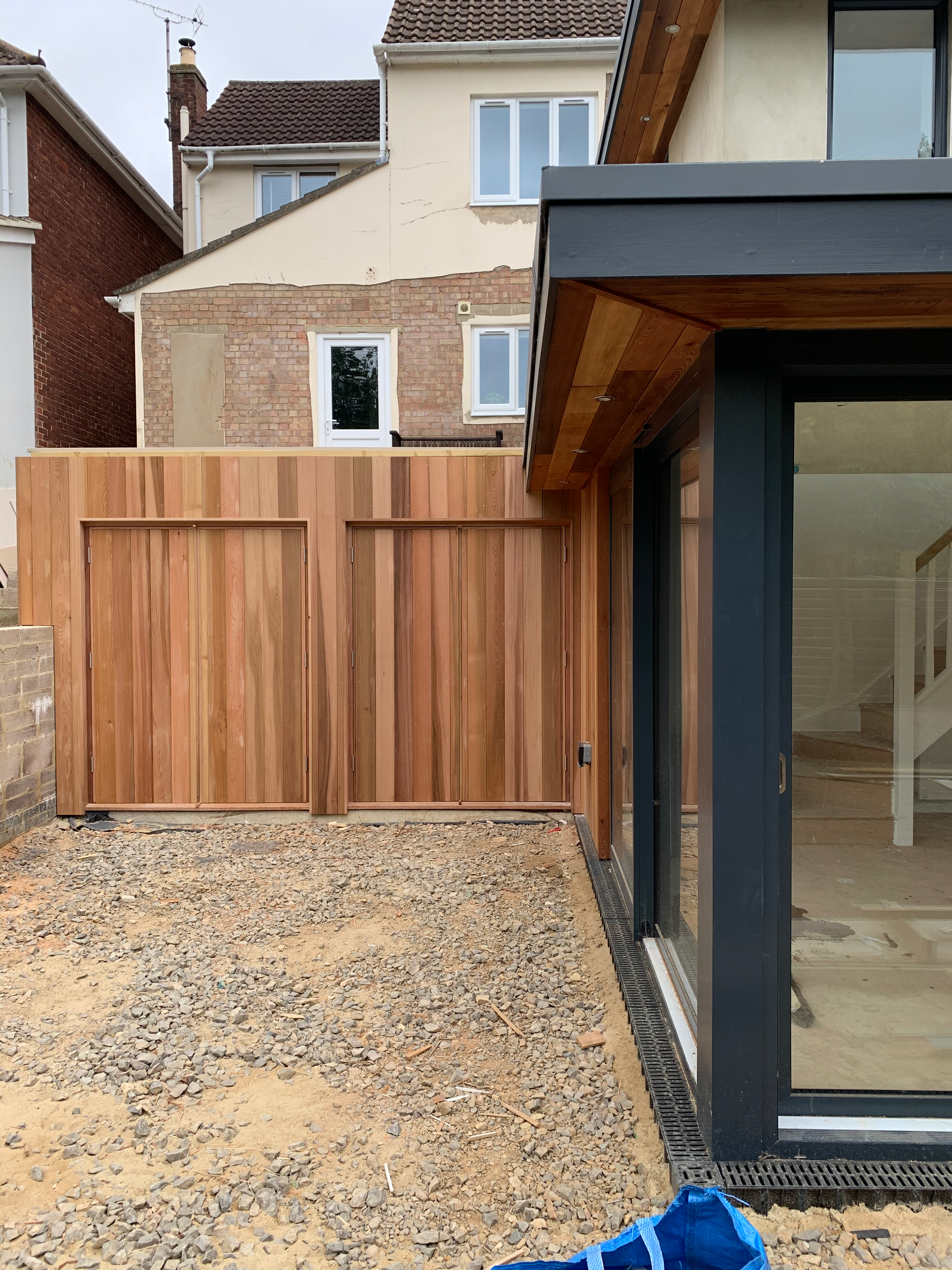 New wooden fence with a gate being installed in a backyard next to a glass door. Gravel covers the ground. Residential house with yellow walls and brick accents is visible in the background.
