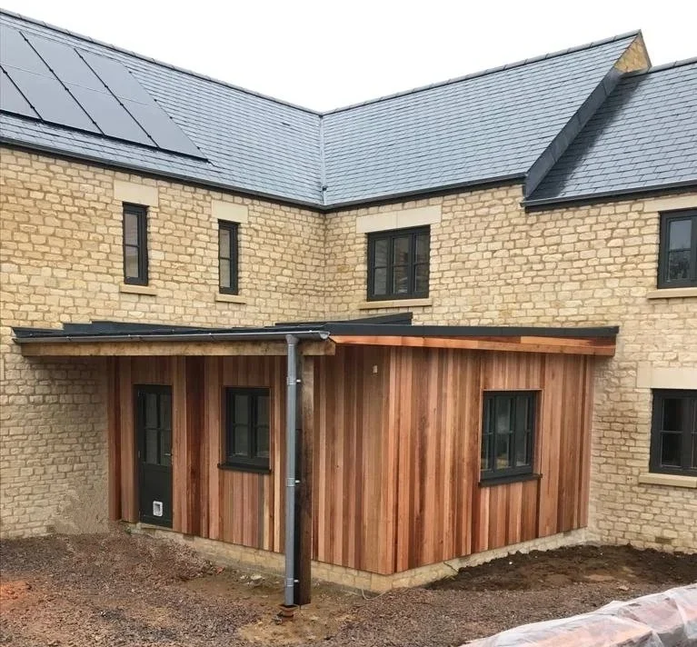 A partially built extension with wooden siding attached to a yellow brick house, featuring black-framed windows and a sloped roof, on an overcast day.