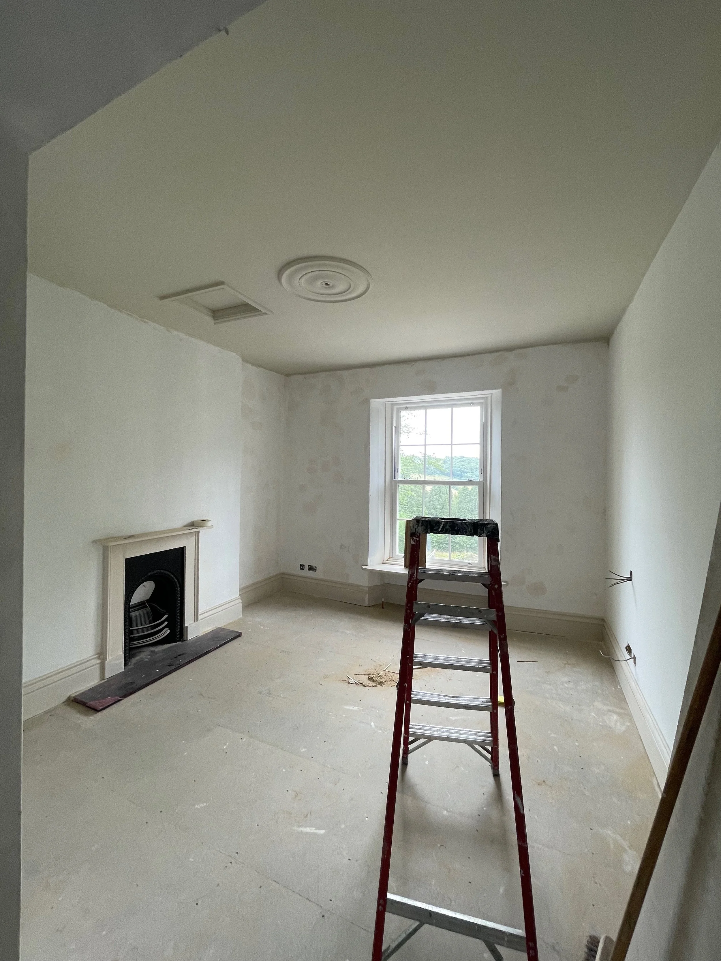 An unfinished living room with a window, a fireplace, and a ladder, indicating ongoing renovation or painting.
