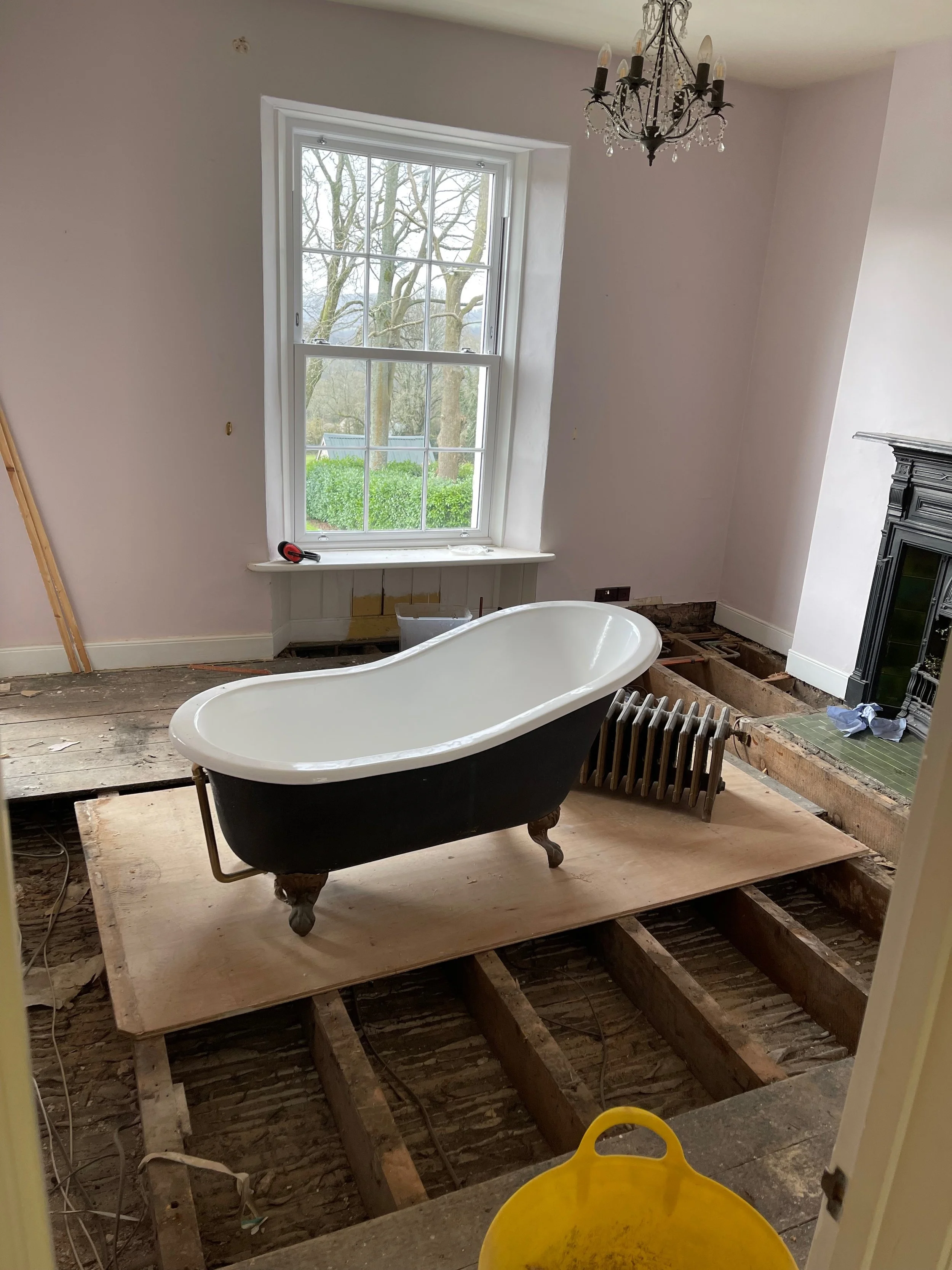 A bathroom under renovation with a clawfoot bathtub placed on a wooden platform, exposed floor framing, a window with a view of trees outside, and a black fireplace surround.