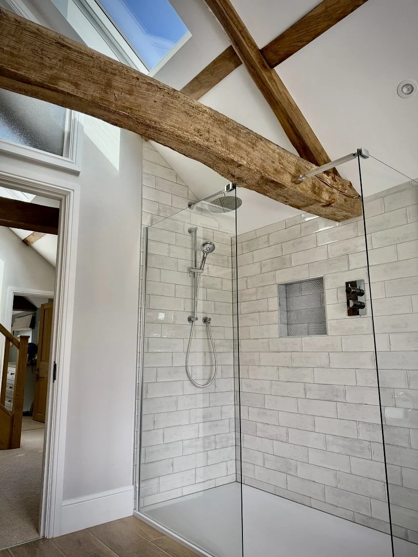 Modern bathroom shower with glass enclosure, white subway tiles, wooden beams on ceiling, small recessed niche with black and white mosaic tile, overhead rain shower, handheld showerhead, and a window.
