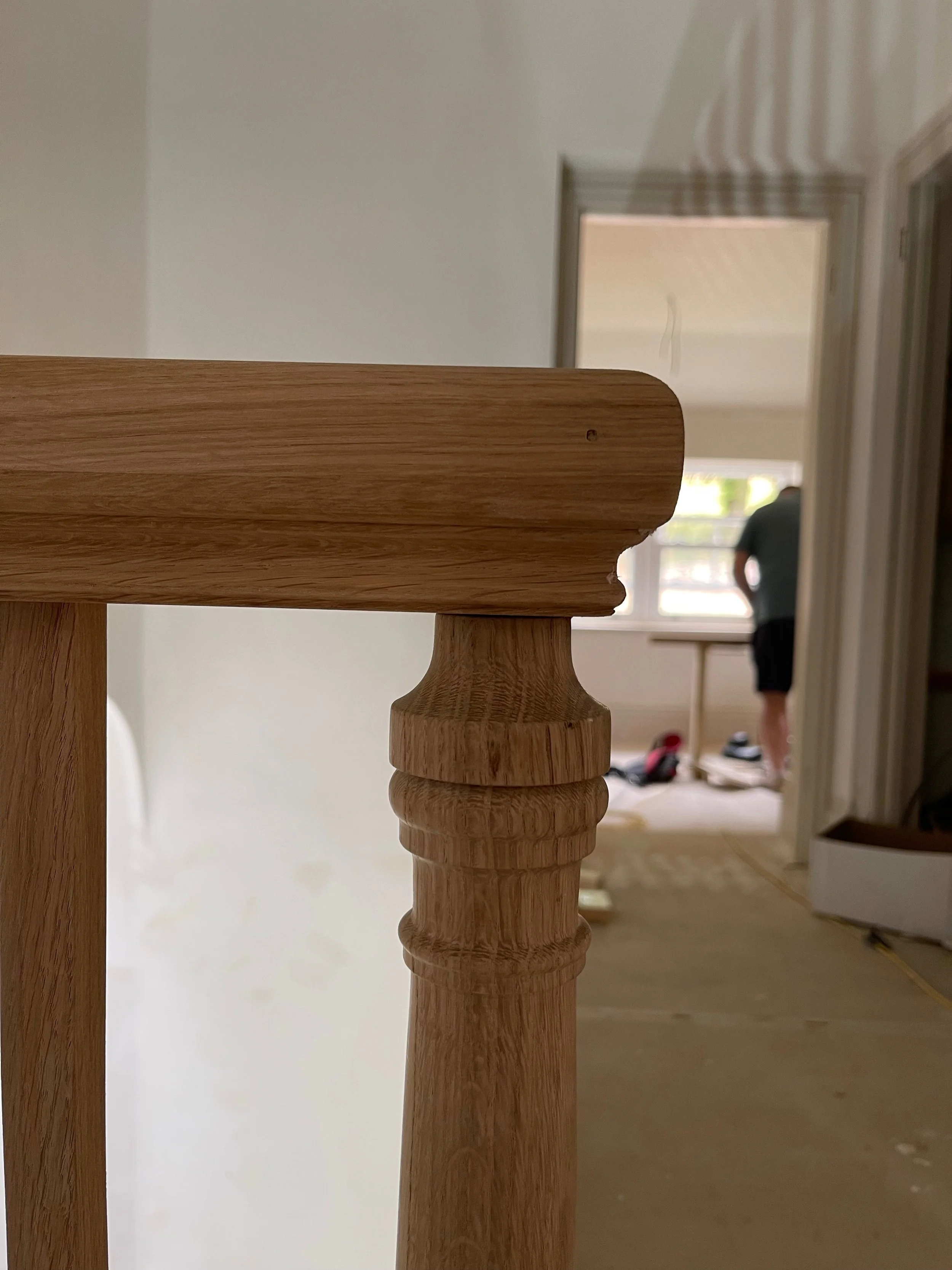 Close-up of a wooden staircase railing with a blurred background showing a person standing near a window in an indoor space under construction or renovation.