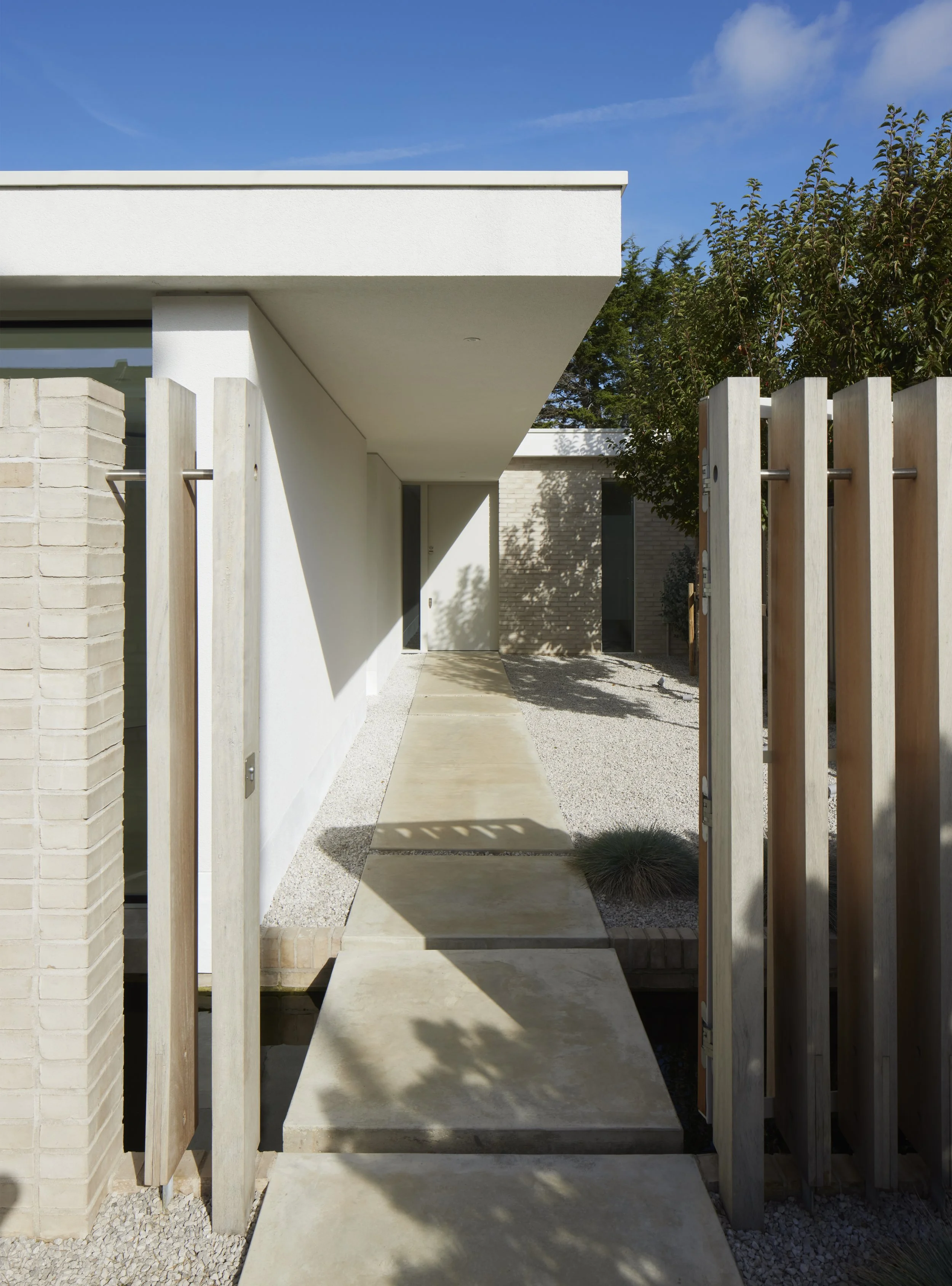 Modern house entrance with a concrete walkway, wooden gate on the right, white exterior walls, and clear blue sky.