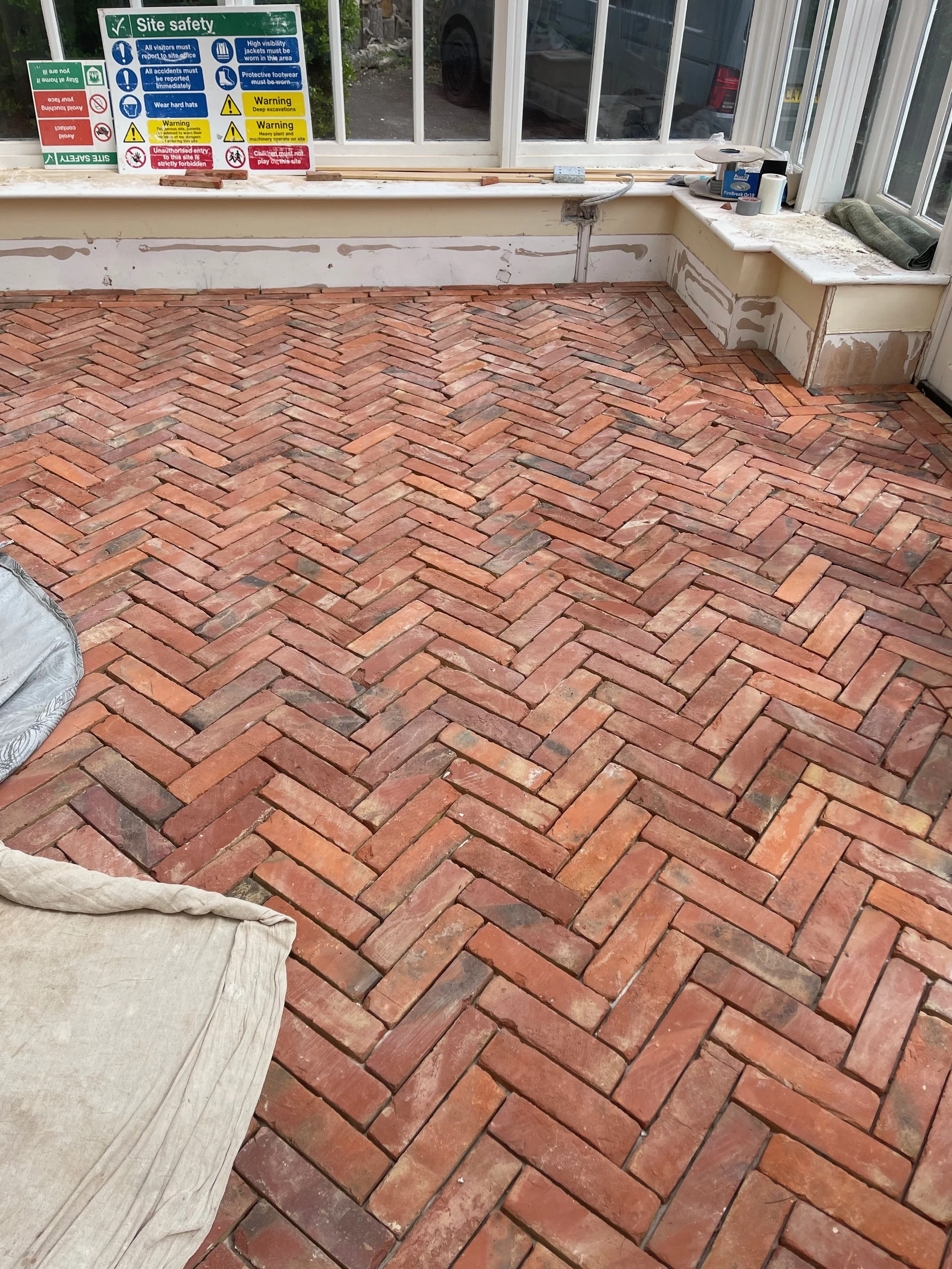 Interior space with newly installed brick herringbone patterned floor, partially painted or repaired walls, windows, and various construction tools and supplies along the window sill.