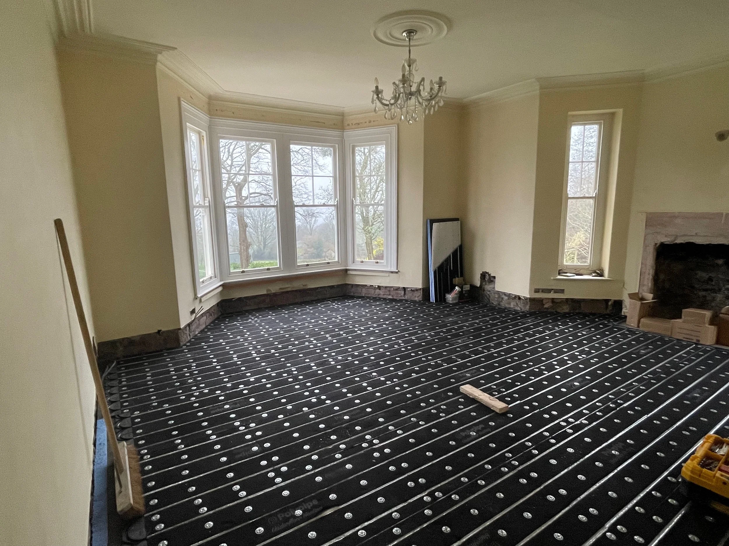 Living room under renovation with black floor heating cables and a large bay window, chandelier, and fireplace.