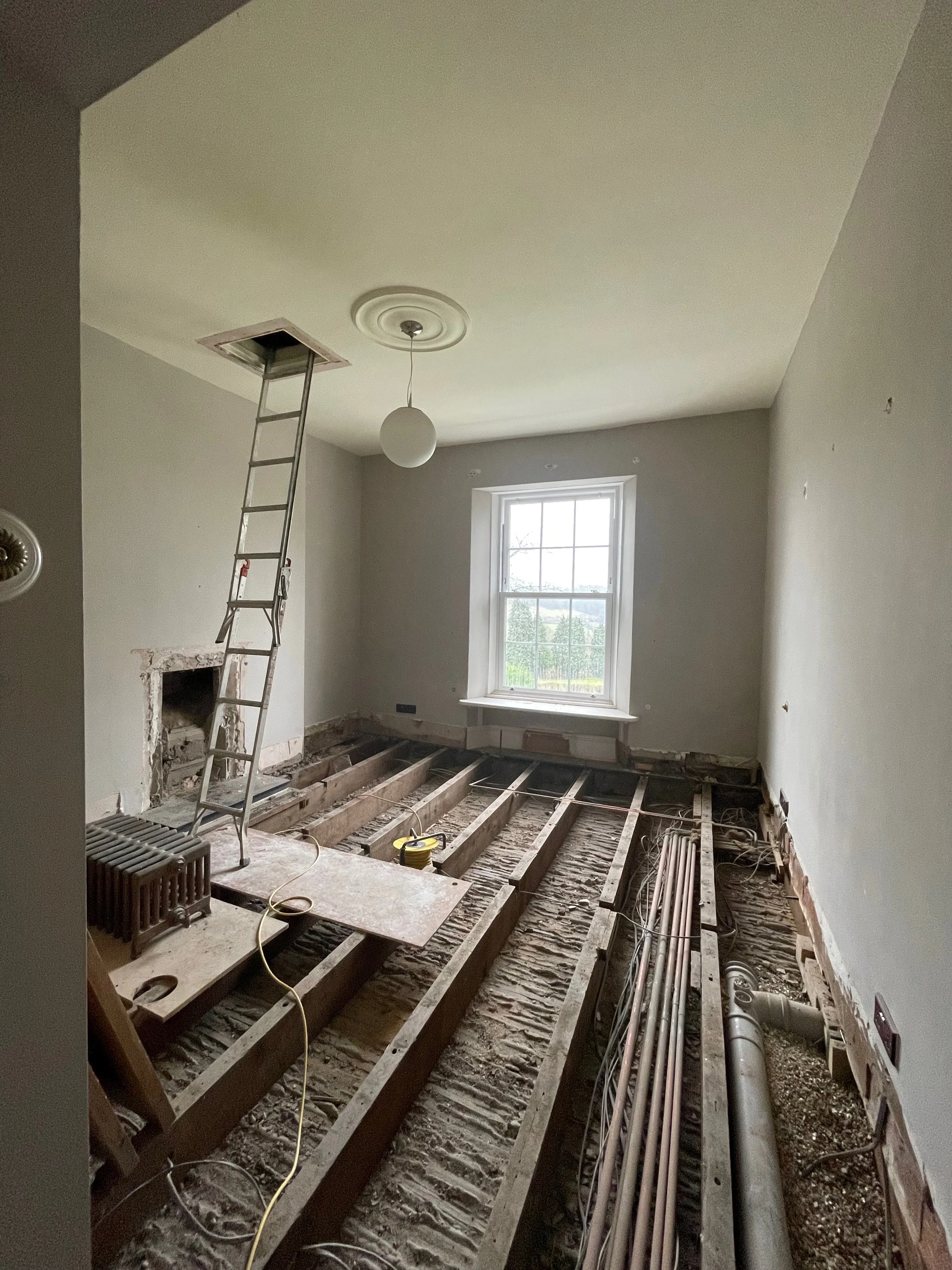 Room under renovation with exposed floor joists, a ladder leaning into the ceiling, a window, and construction tools and materials.