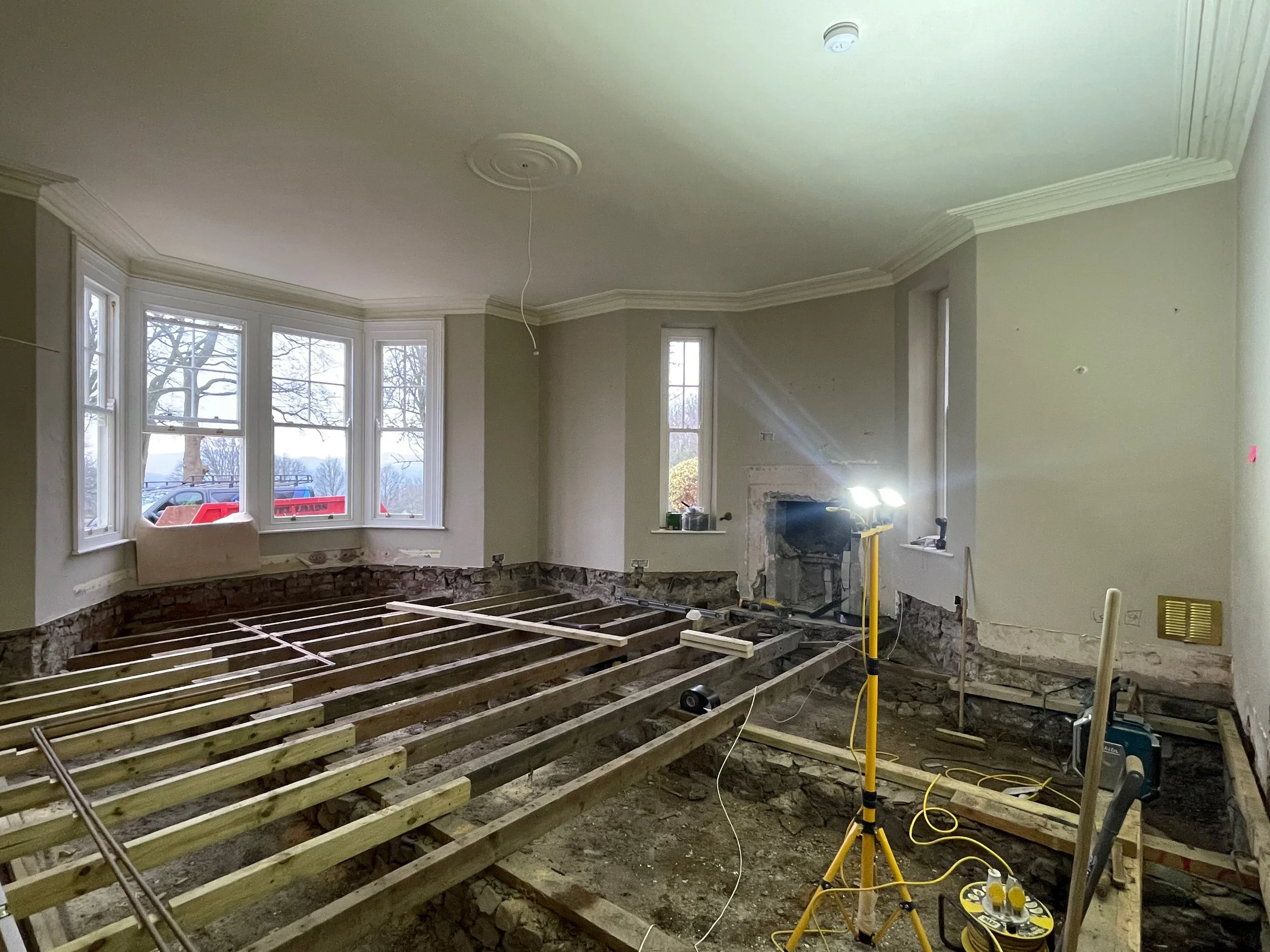 Living room under renovation with exposed wooden floor framing, construction equipment, and scaffolding, with large windows.
