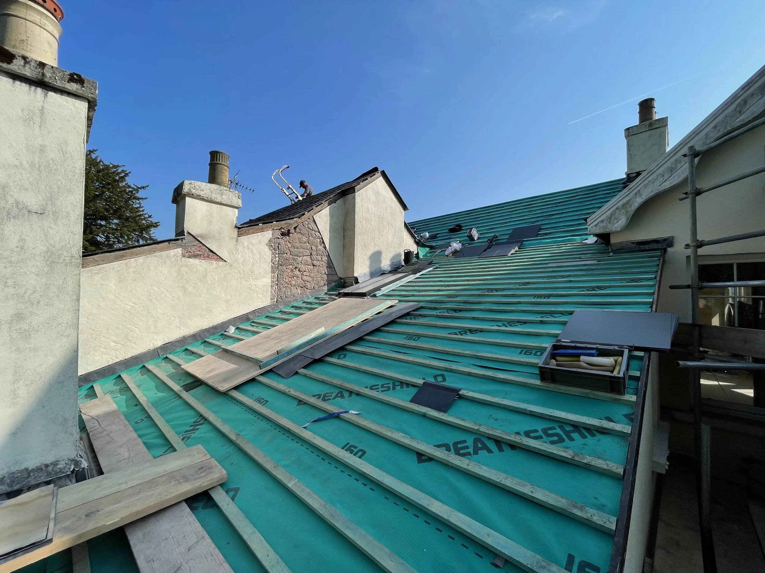 Construction workers installing new roofing on a house with green underlayment and wooden battens, blue sky in background.
