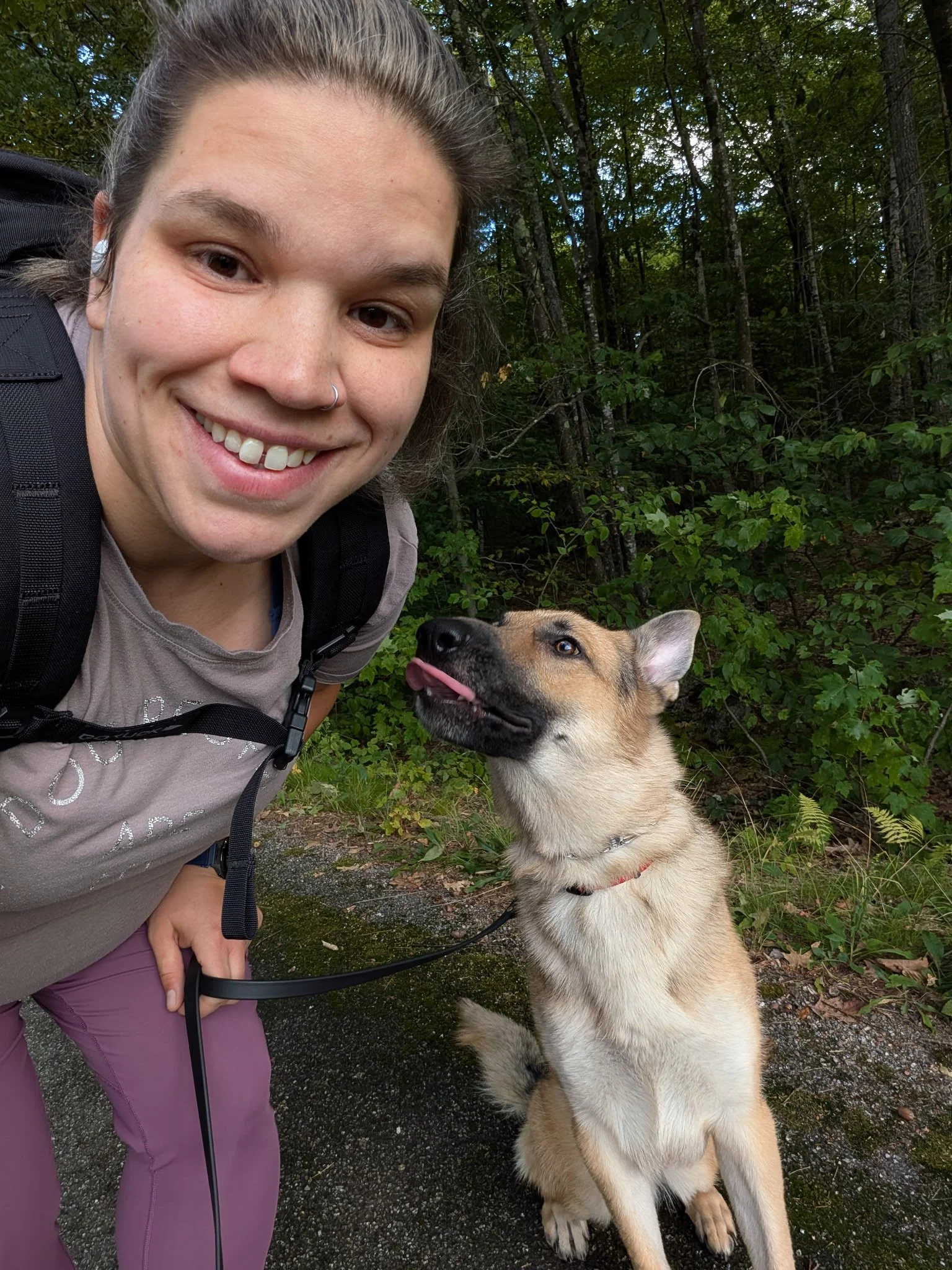 A young woman smiling outdoors with a dog sitting beside her on a forest trail.