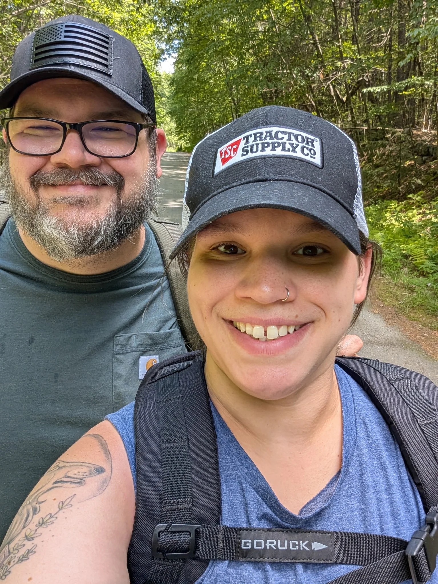 Two smiling women outdoors on a forest trail, wearing caps and backpacks.