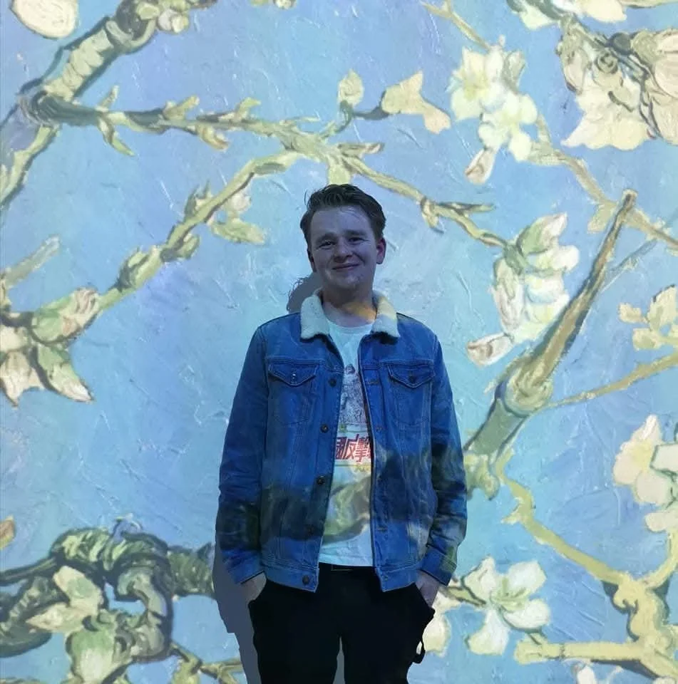 A young man wearing a denim jacket and a graphic T-shirt stands in front of a projected painting of flowering branches on a light blue wall.