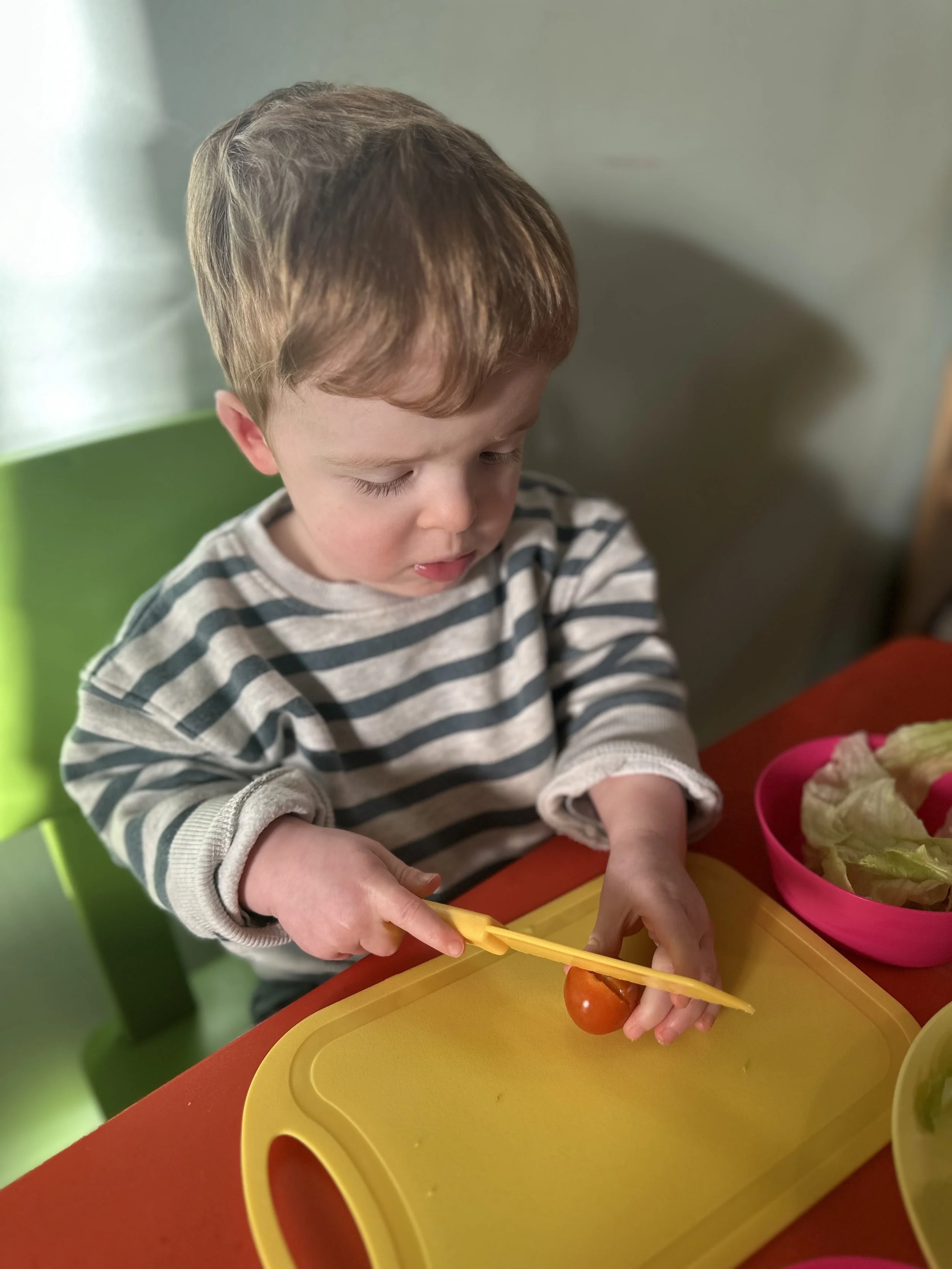 A young boy with brown hair wearing a striped shirt is sitting at a table, slicing a cherry tomato on a yellow cutting board with a plastic knife.