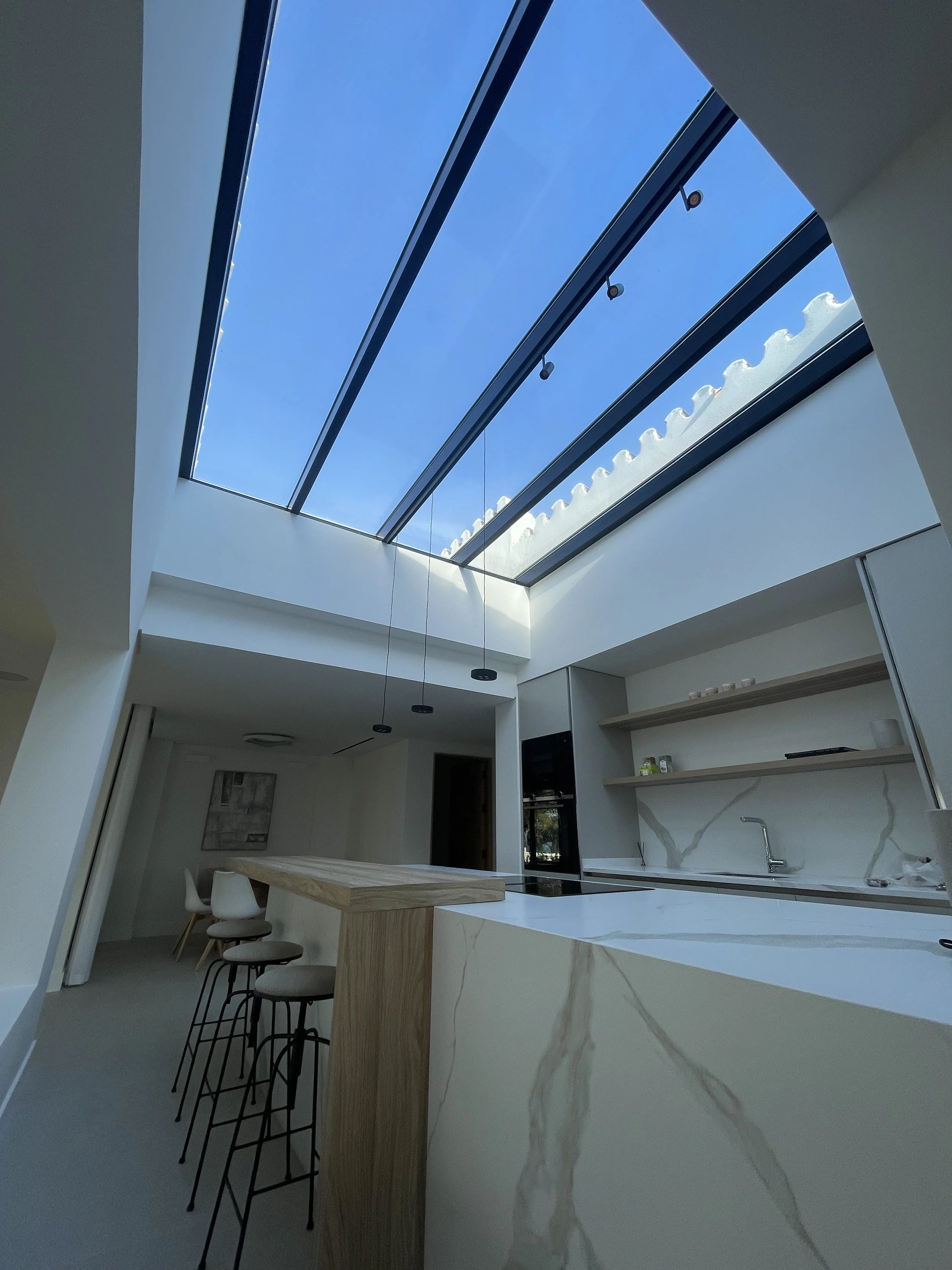 Cocina moderna con techo de claraboya grande que permite la entrada de luz natural, encimera de mármol y barra de madera con sillas.