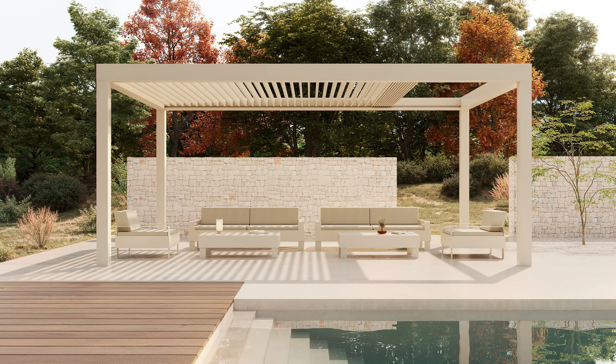 Modern outdoor patio with white furniture, including chairs, a sofa, and coffee tables, under a pergola, with a swimming pool and a stone wall in the background, surrounded by trees with autumn foliage.