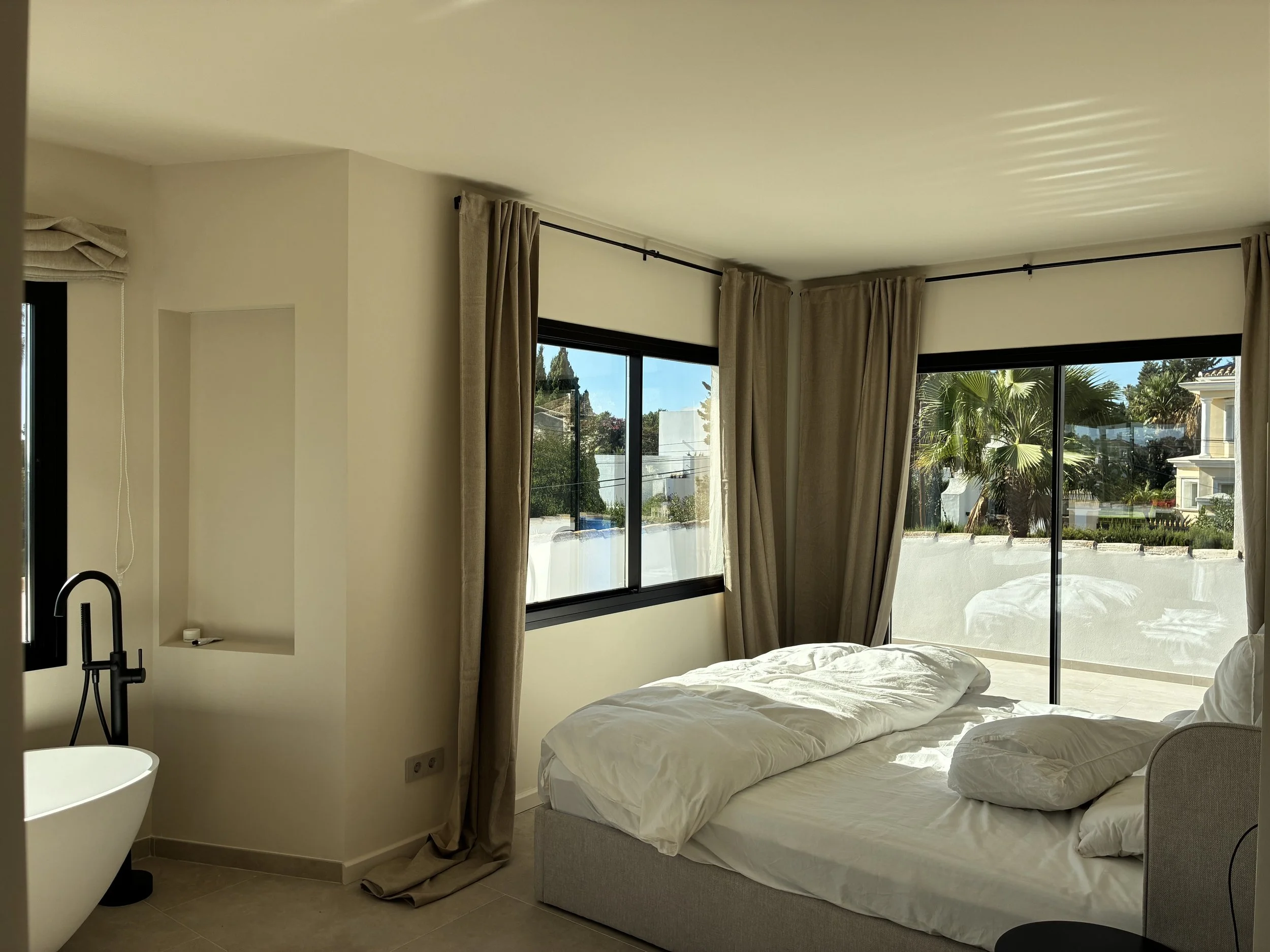 Habitación con cama desordenada, ventanas grandes con cortinas beige, vista al exterior con plantas y casas, y un lavabo en la esquina izquierda.