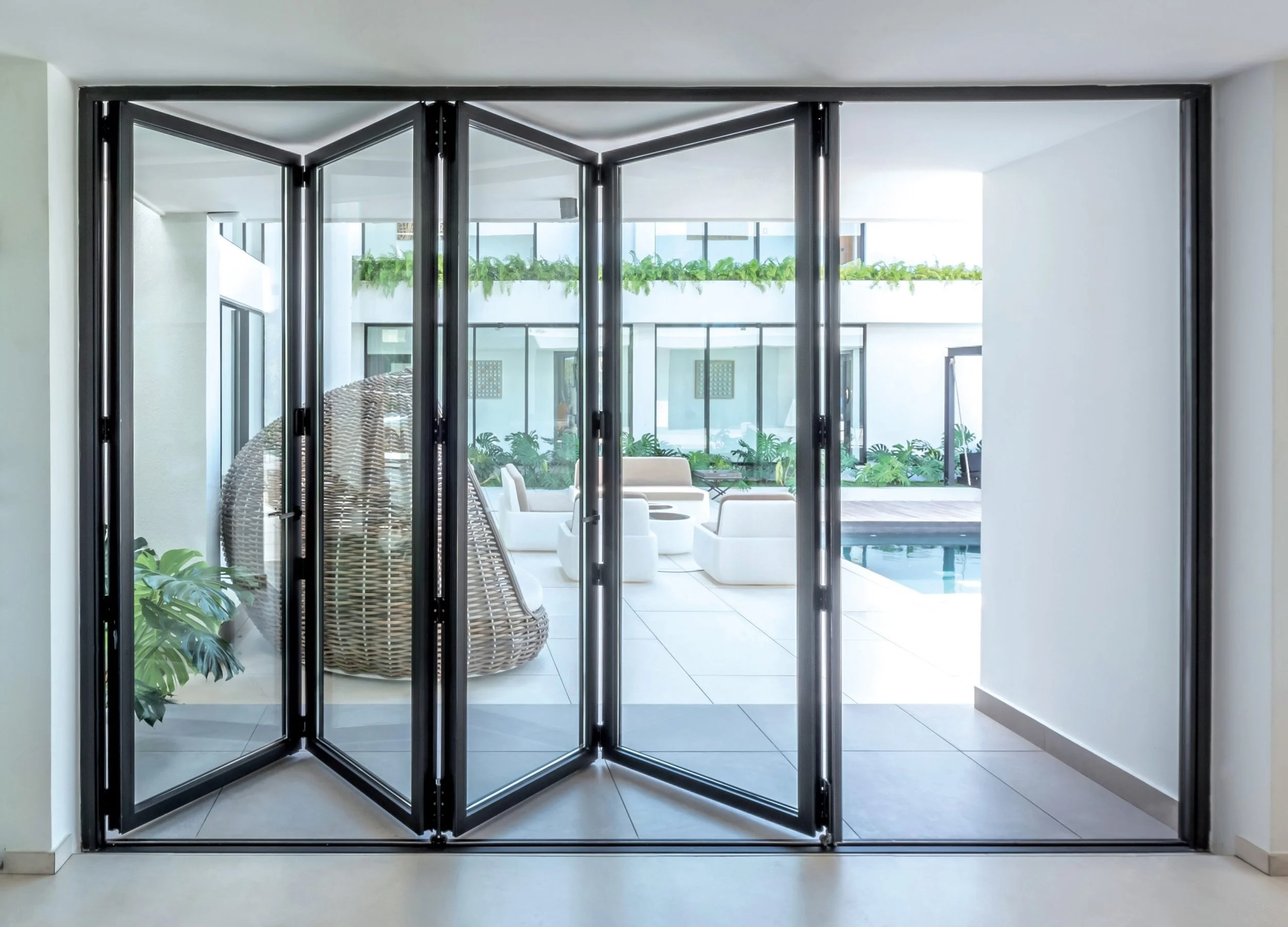 Vista de un patio con sillones blancos y área de alberca, vista desde dentro de una casa mediante puertas corredizas de vidrio y marco negro.