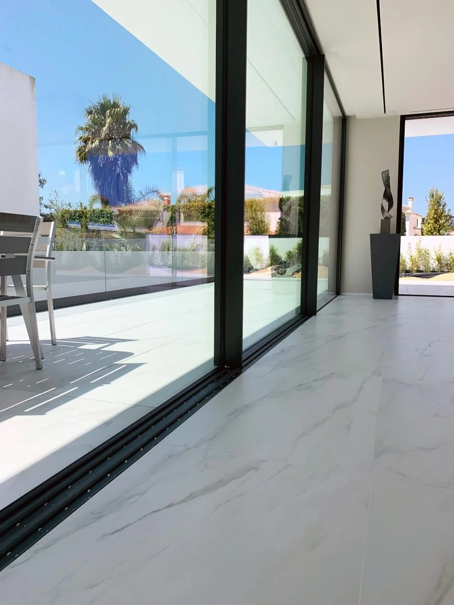 Vista desde el interior de una sala moderna con piso de mármol y grandes ventanas de vidrio que llevan a una terraza exterior con muebles y vegetación.